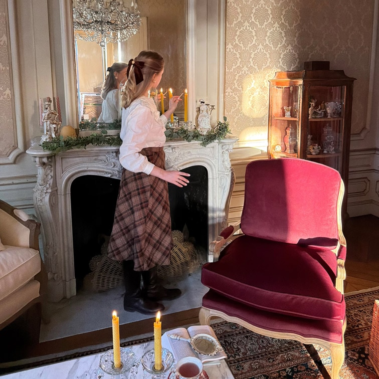 Mujer encendiendo velas junto a chimenea de mármol en salón elegante con sillón burdeos