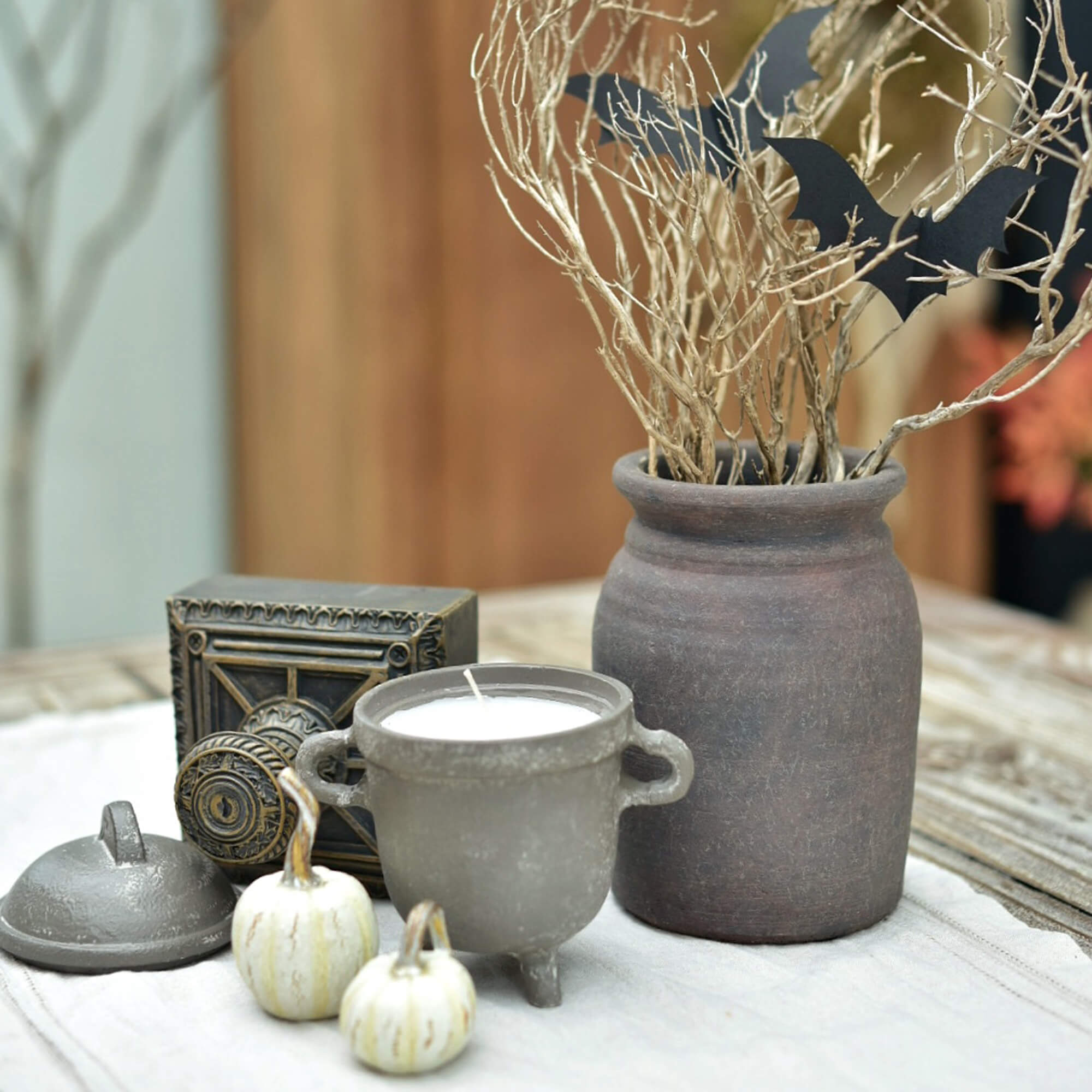 Conjunto decorativo con vela de cerámica, jarrón de barro y pequeñas calabazas blancas sobre mesa