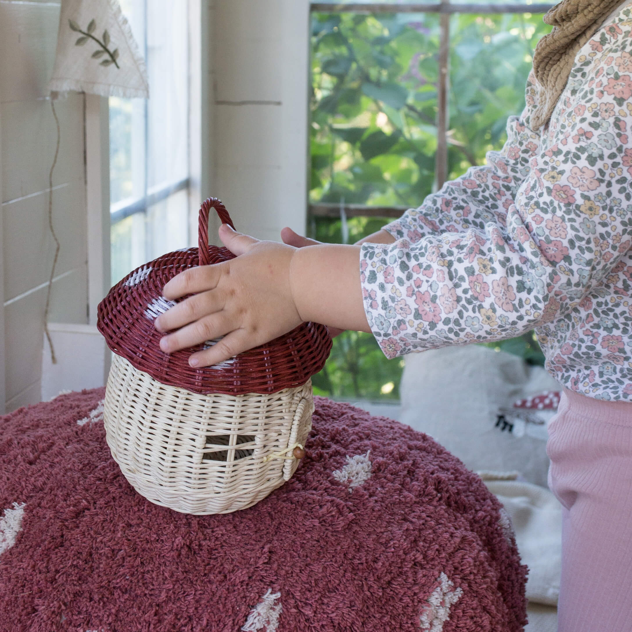 Niño tocando cesta de mimbre en forma de seta beige y roja sobre alfombra rosa