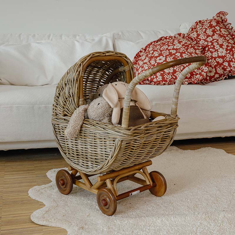Cochecito infantil de mimbre con ruedas de madera y peluches en sala luminosa con sofá blanco