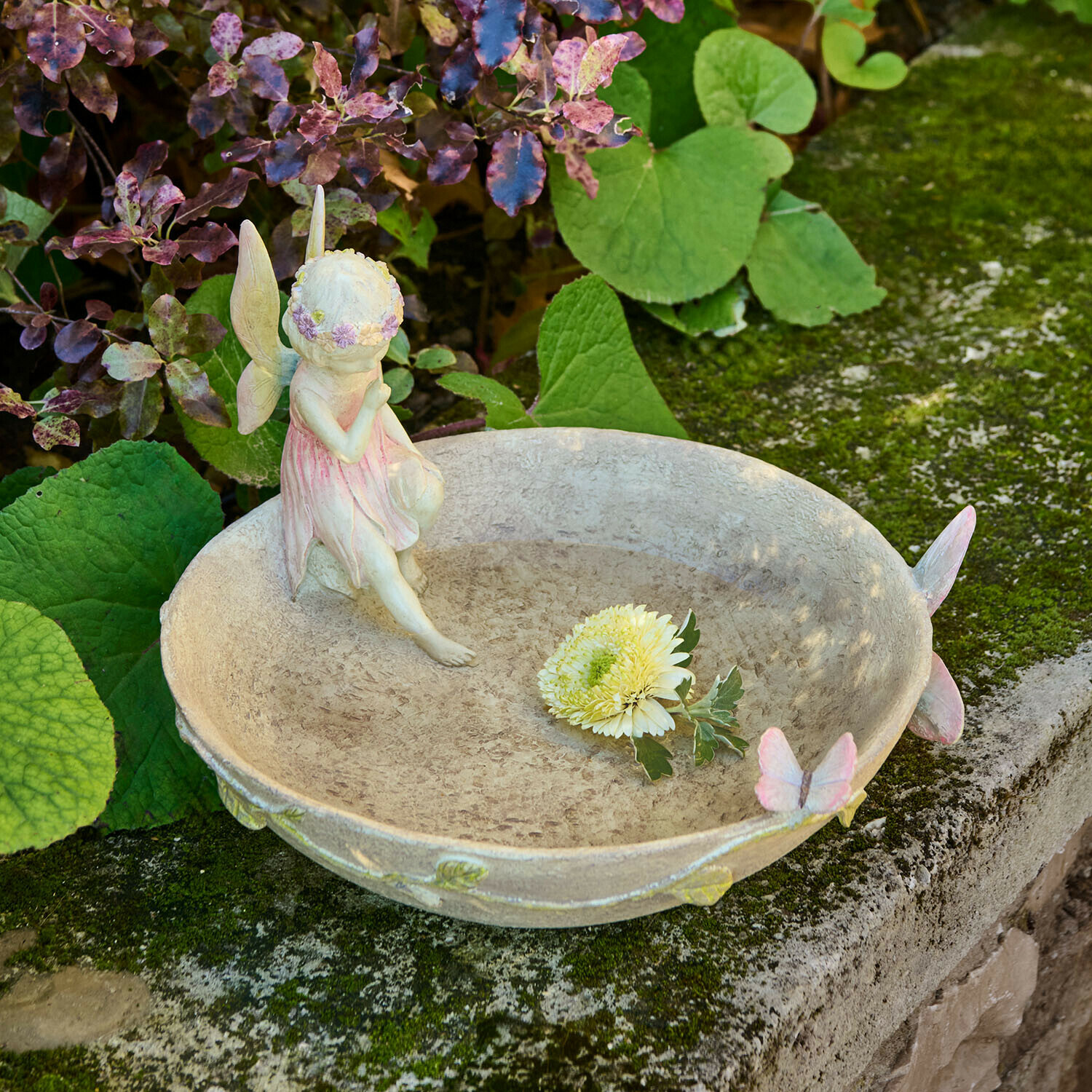 Cuenco decorativo de jardín con figura de hada y mariposas en colores pastel sobre muro de piedra