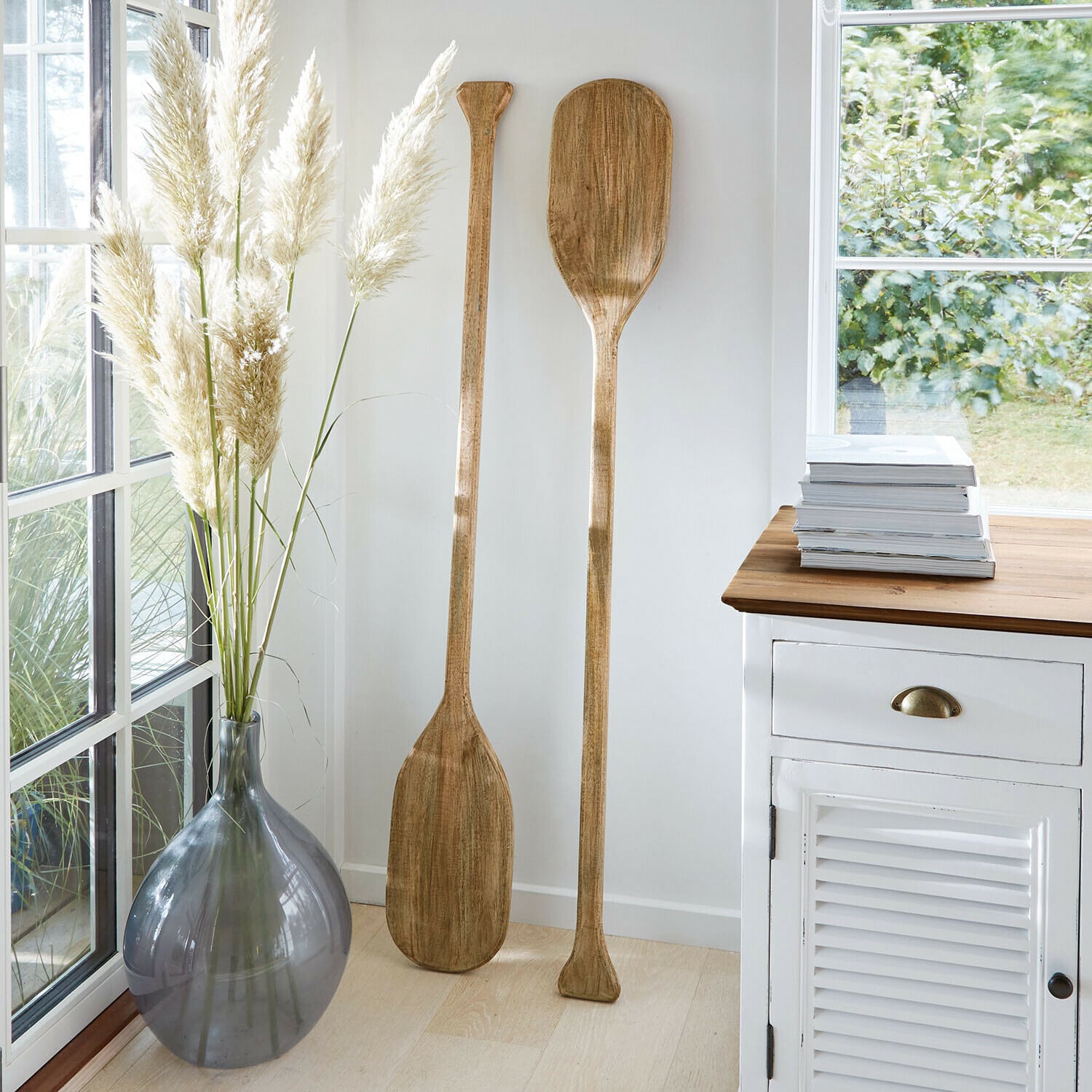 Set de 2 remos decorativos Windermount Dos remos decorativos de madera en interior luminoso junto a jarrón gris y mueble blanco