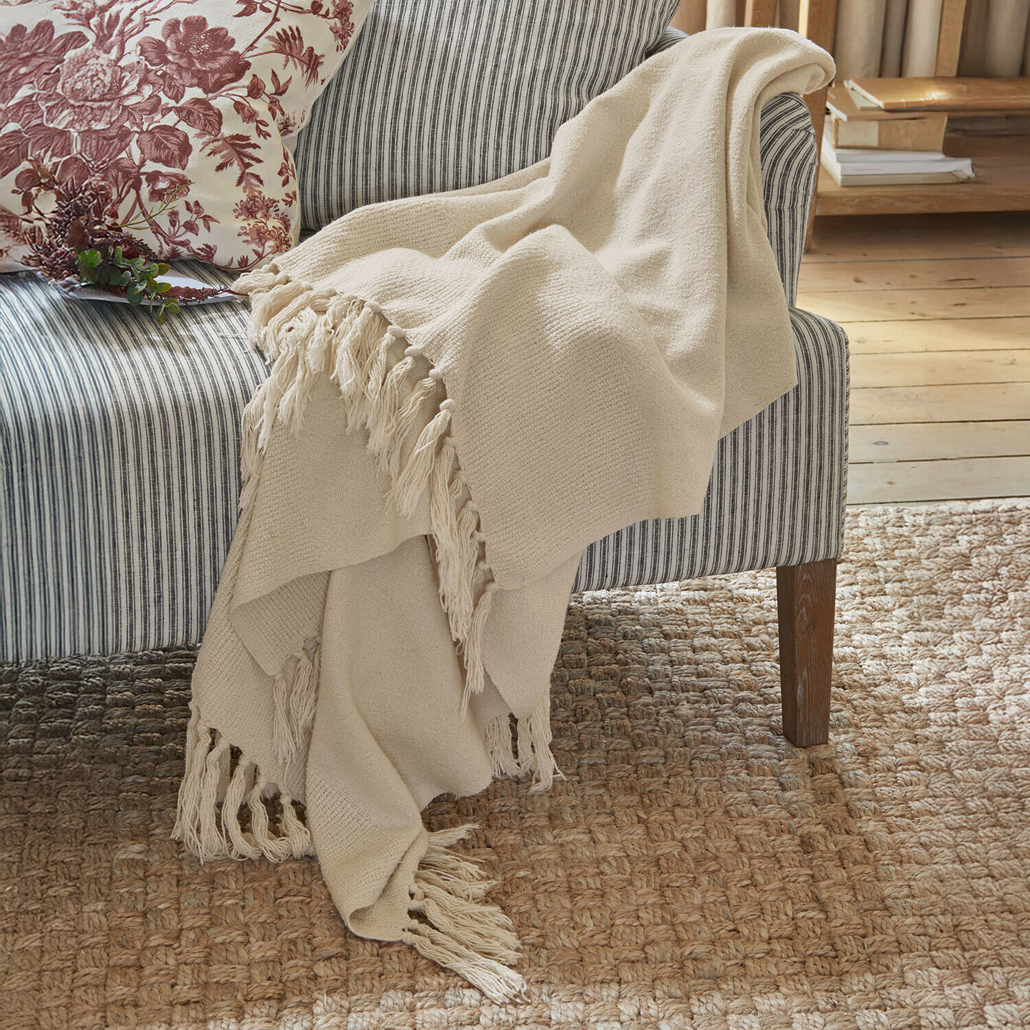 Plaid Skandira Manta beige con flecos de tela suave sobre un sofá gris a rayas en un ambiente acogedor