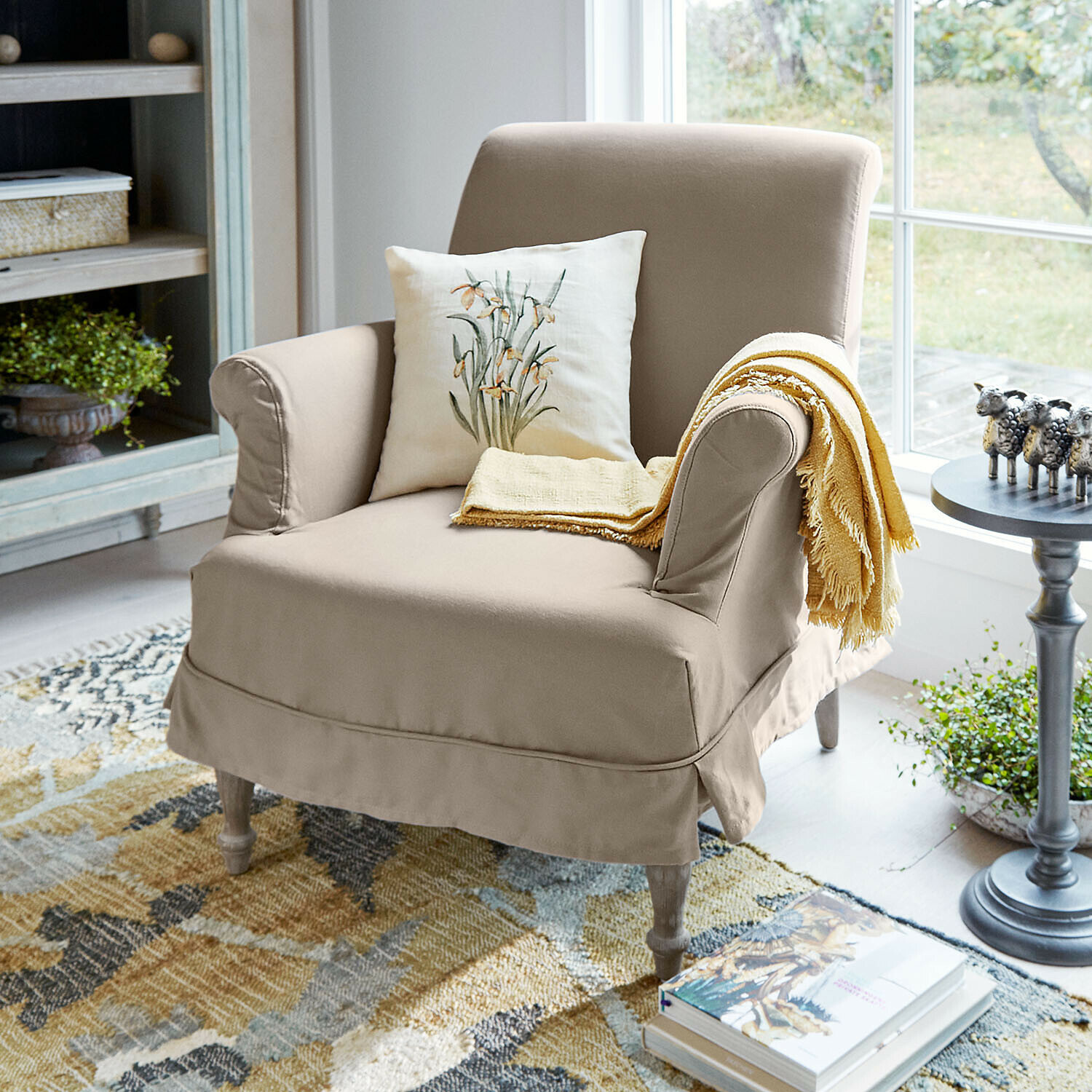 Sillón Everleighs Sillón beige de tela con cojín floral y manta amarilla en sala luminosa con alfombra colorida