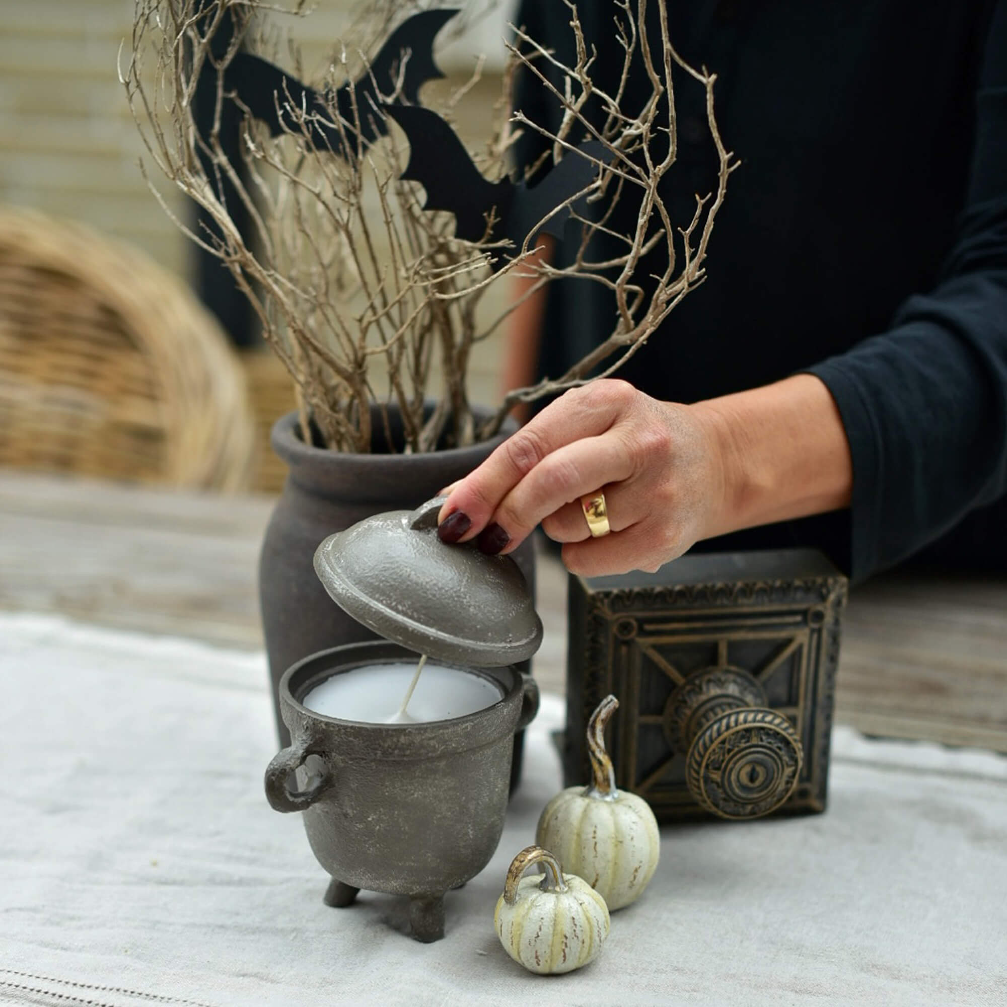 Vela gris en forma de caldero con tapa sobre mesa decorada para Halloween con calabazas y ramas