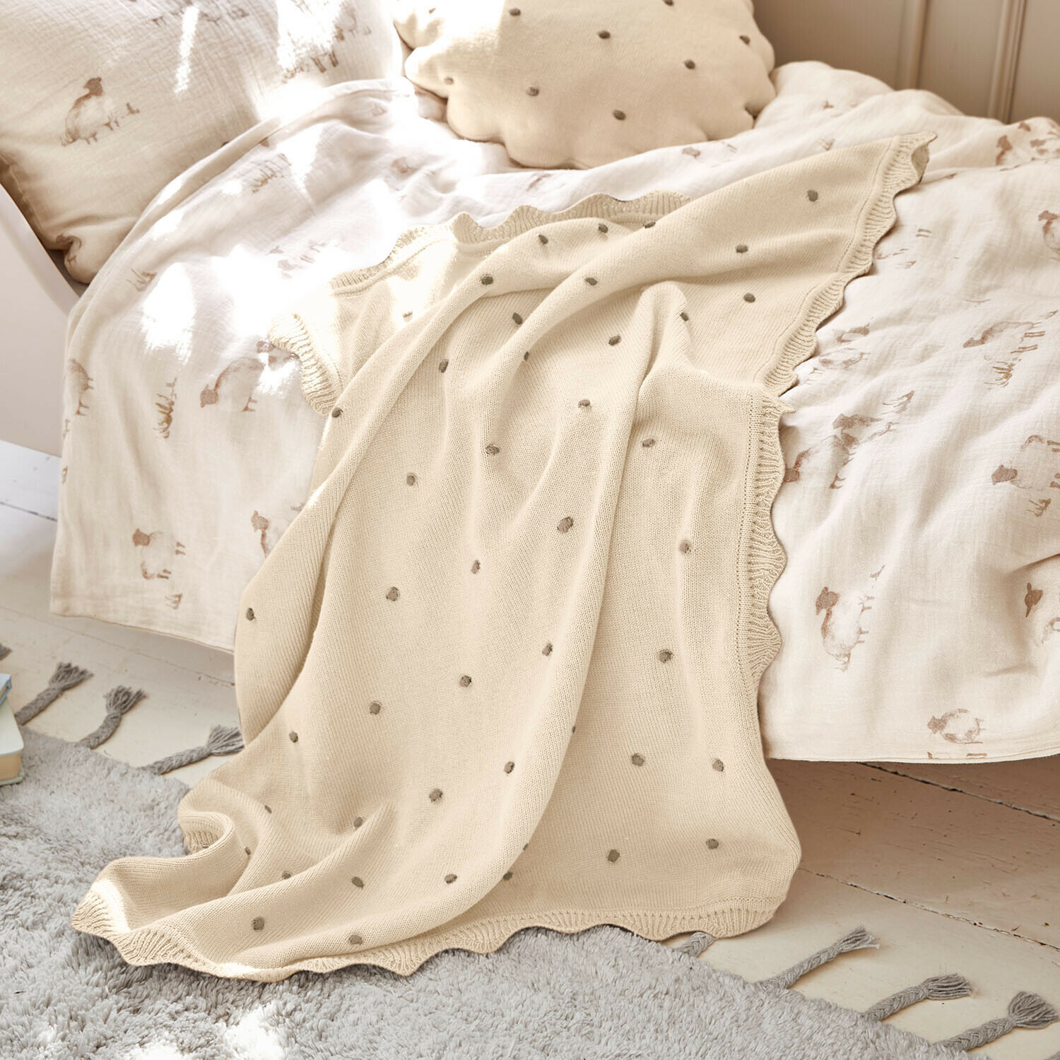 Plaid Saphie Manta de punto beige con lunares grises sobre cama con ropa de cama clara y cojín natural