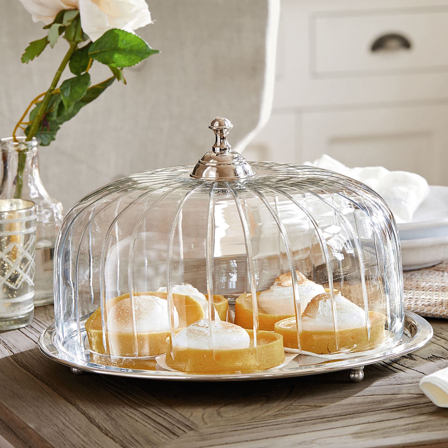 Campana para tarta Allaire Campana de cristal con asa metálica sobre mesa de madera con tartas de limón dentro