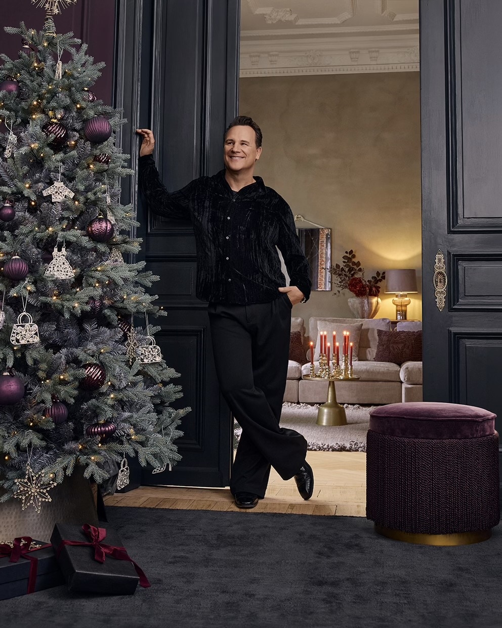 Hombre con ropa negra junto a árbol de Navidad decorado con bolas moradas en salón elegante con velas rojas encendidas