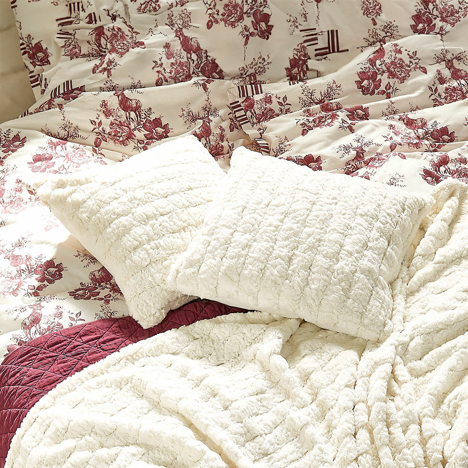 Cojines y mantas de felpa crema sobre cama con ropa de cama con estampado floral y ciervos burdeos