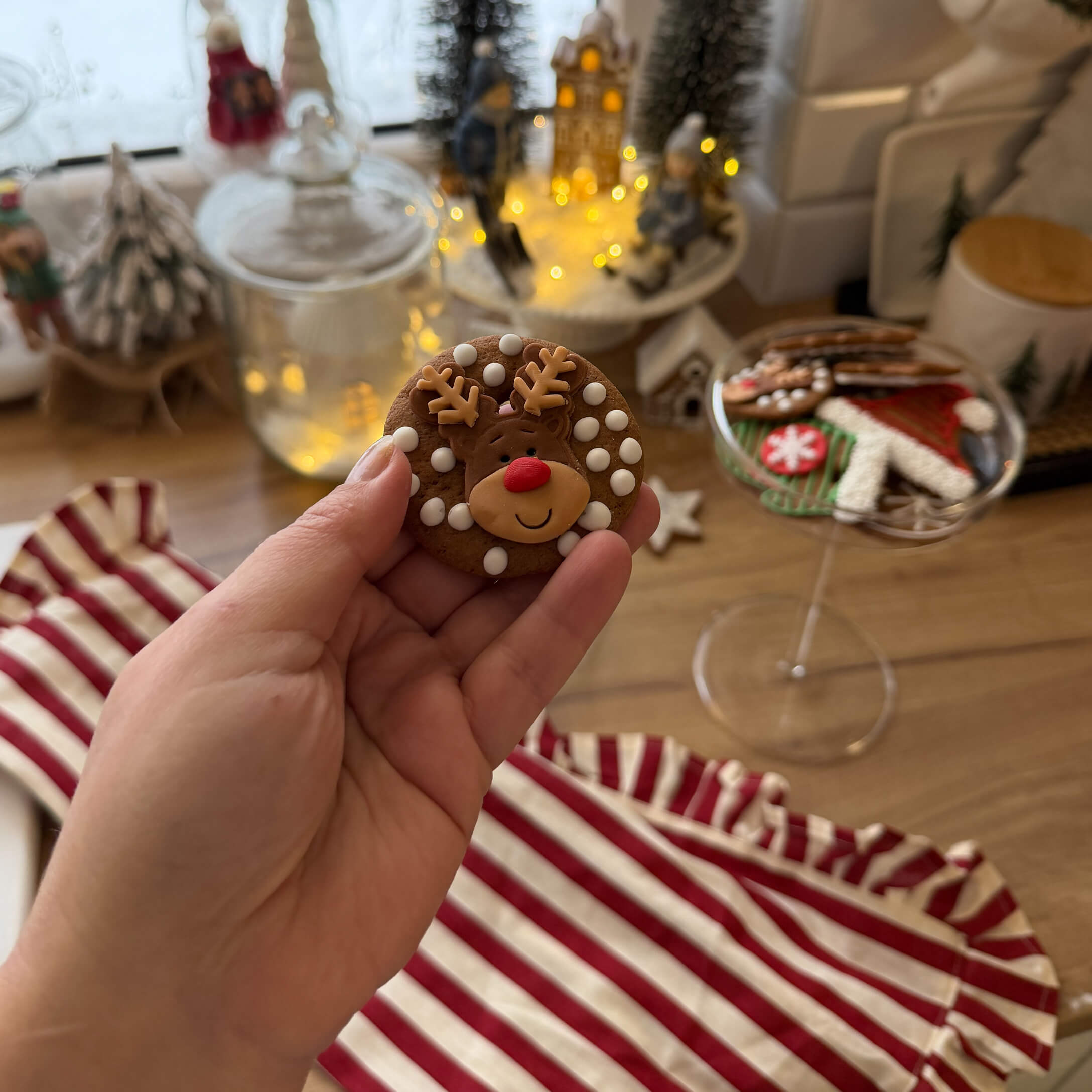 Mano sosteniendo galleta de jengibre con reno y puntos blancos, fondo con decoraciones navideñas y galletas