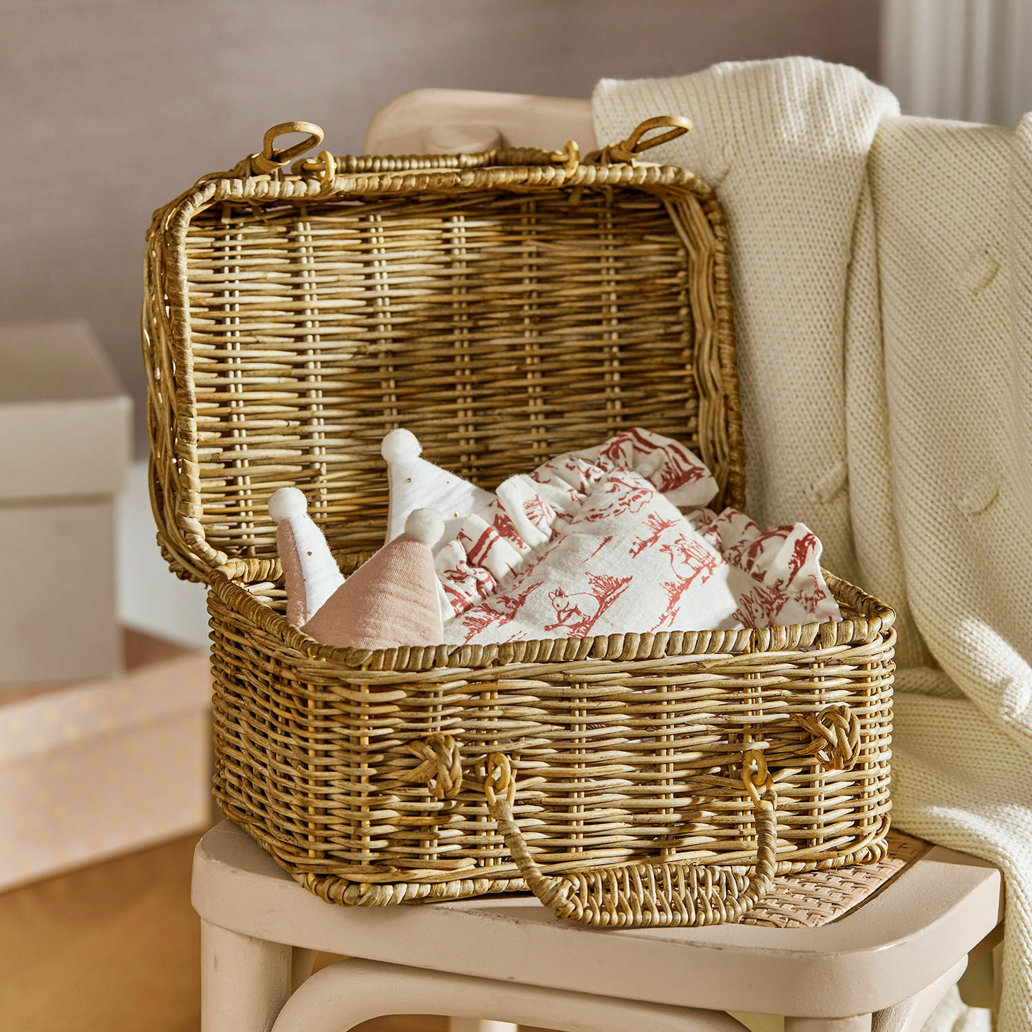 Maleta Fableton Cesta de mimbre con tapa sobre silla, llena de telas con estampados rojos y juguetes suaves