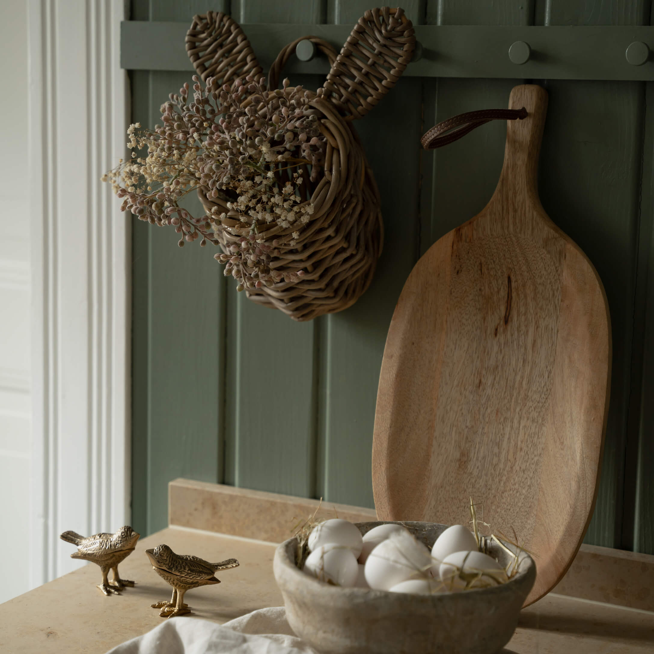 Cesta de mimbre con forma de conejo con flores secas y tabla de cortar de madera sobre fondo verde