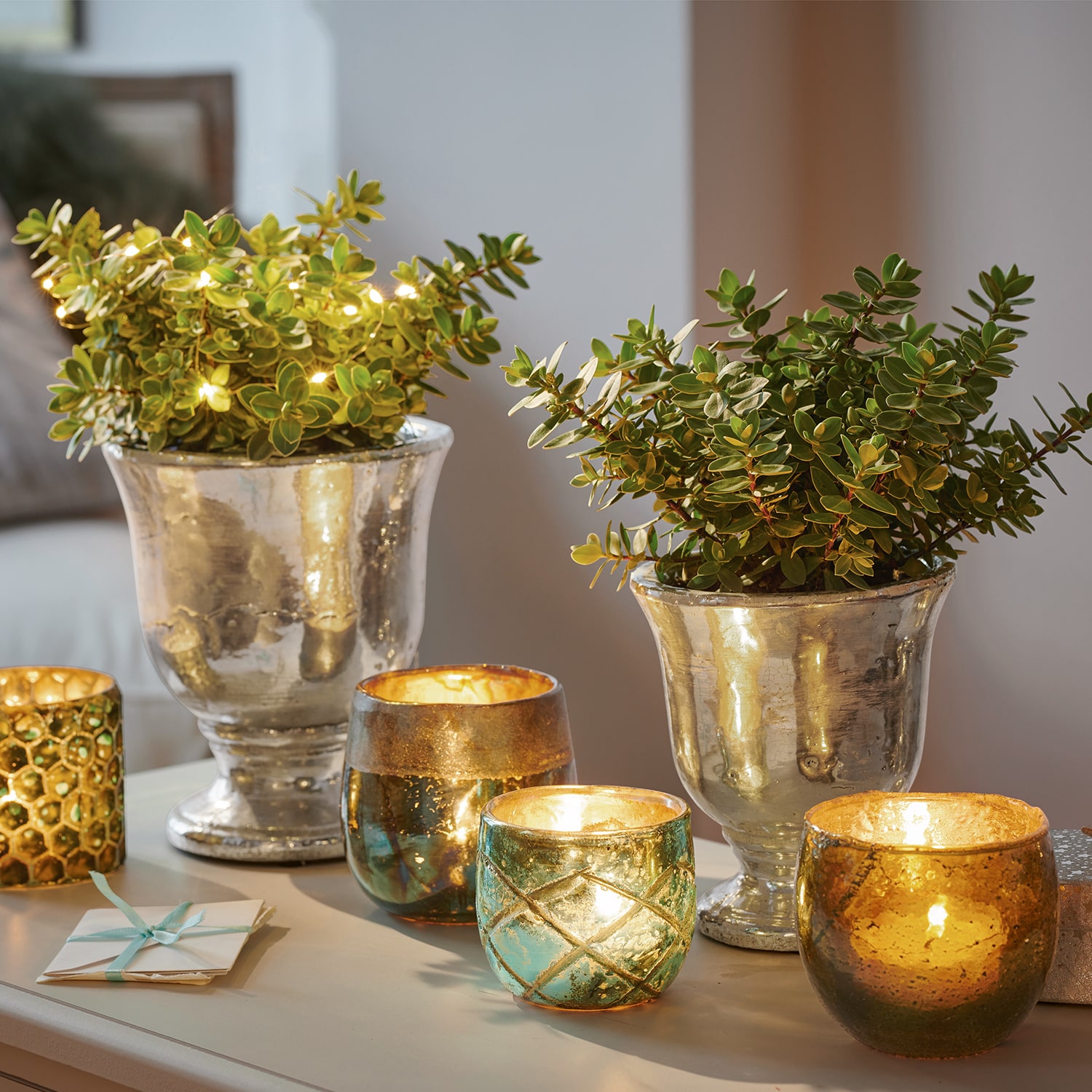 Macetas decorativas plateadas con plantas verdes y portavelas de vidrio iluminados sobre mesa