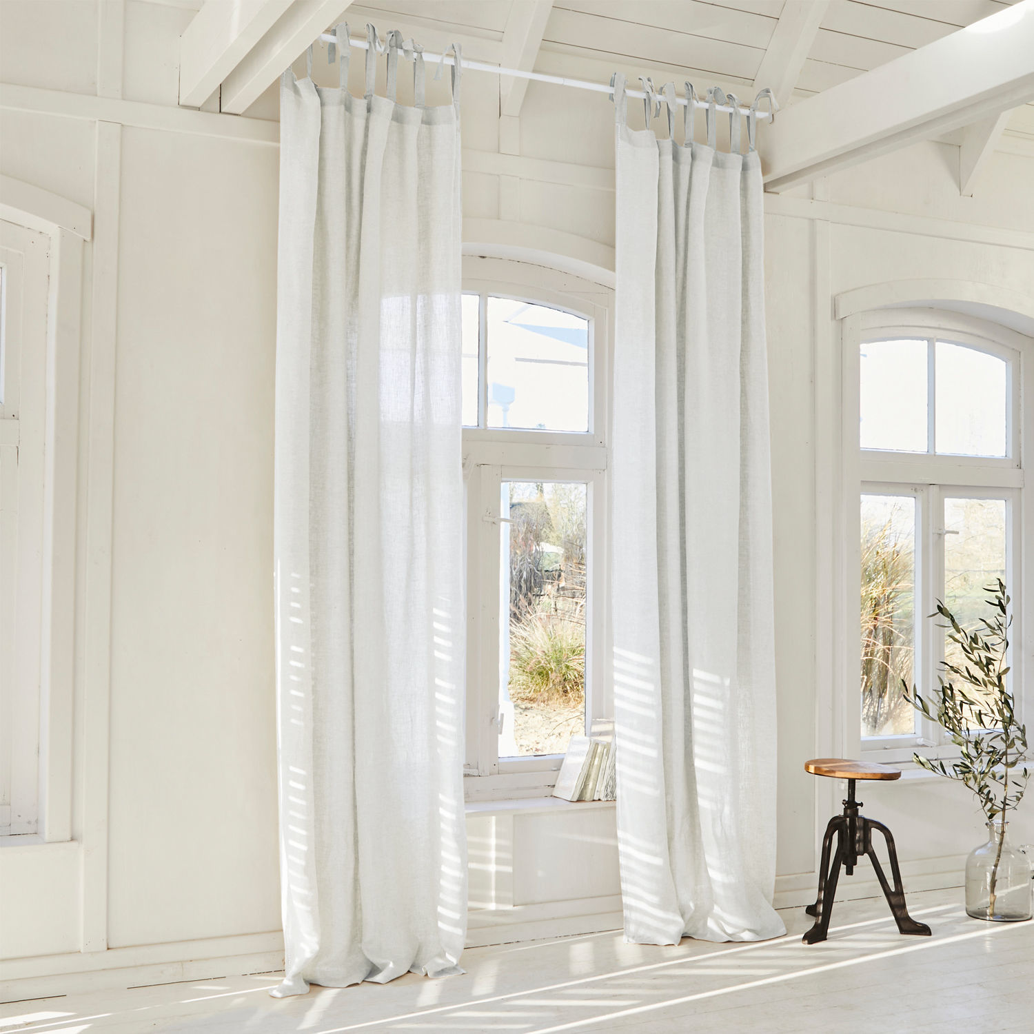 Cortina Monthelie Cortinas blancas de lino colgadas en habitación luminosa con suelo de madera y taburete