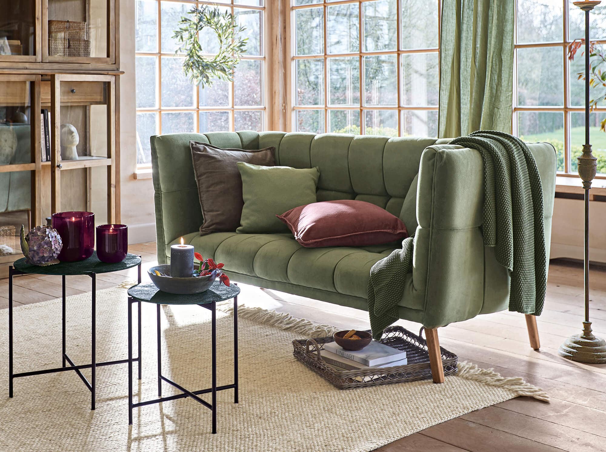 Sofá vintage verde de terciopelo con cojines y manta en salón luminoso con grandes ventanas