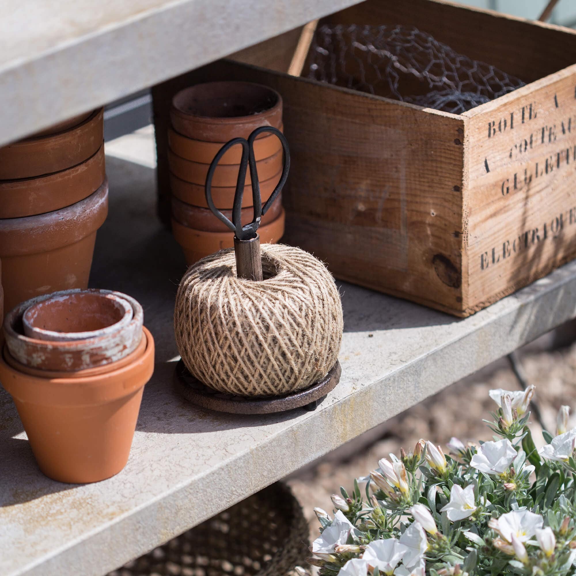Hilo natural de jardinería con tijeras de metal sobre estante de cemento junto a macetas de terracota y caja de madera