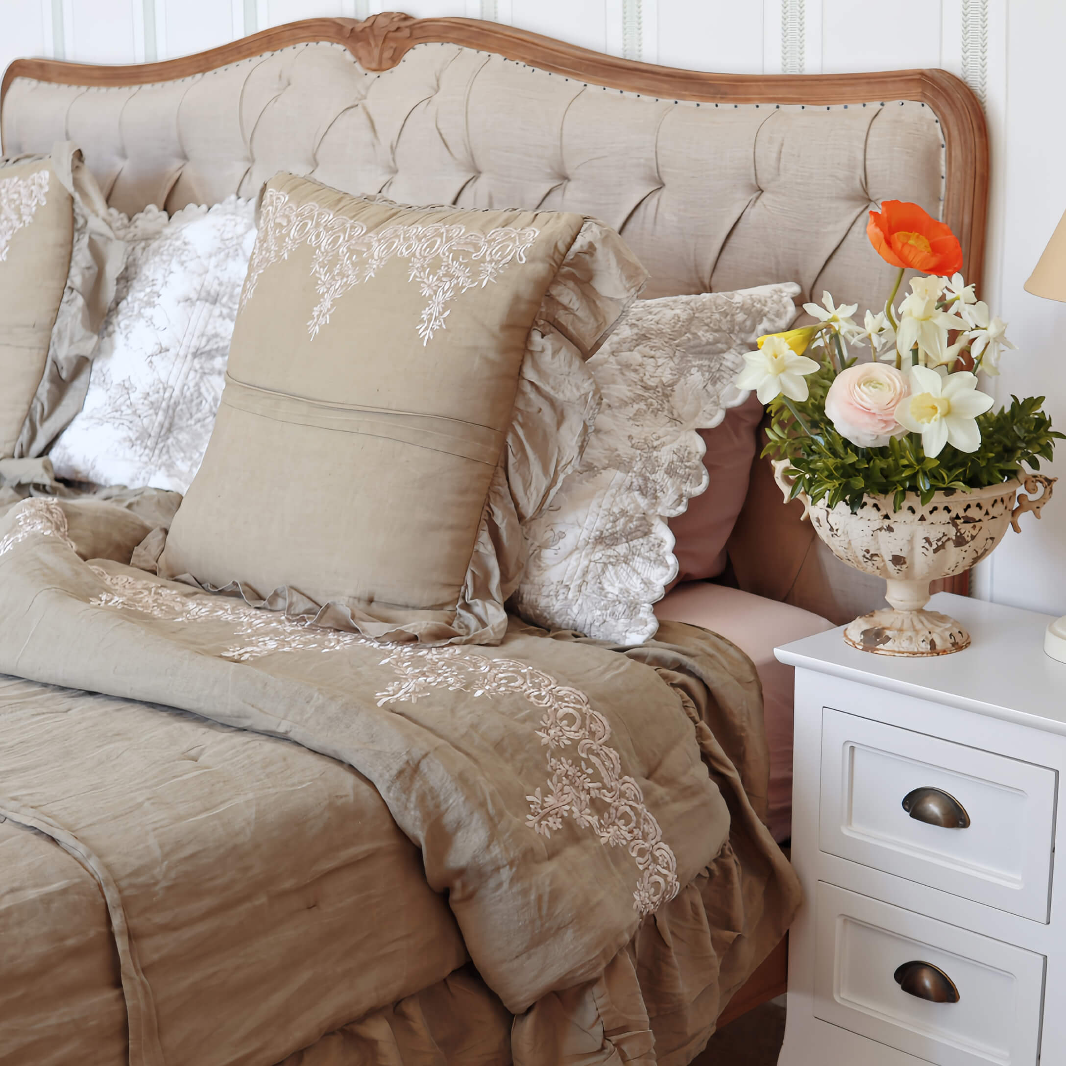 Ropa de cama beige con cojines bordados y colcha sobre cama con cabecero tapizado