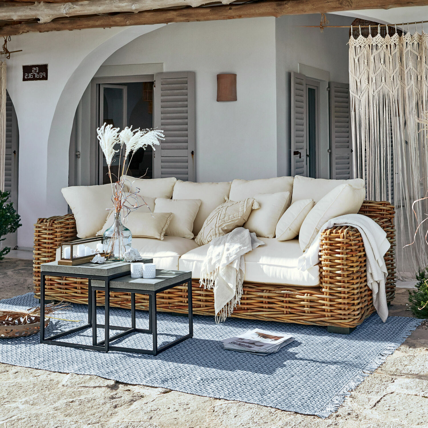 Sofa Rimini Sofá de ratán con cojines crema y manta sobre alfombra azul en terraza soleada