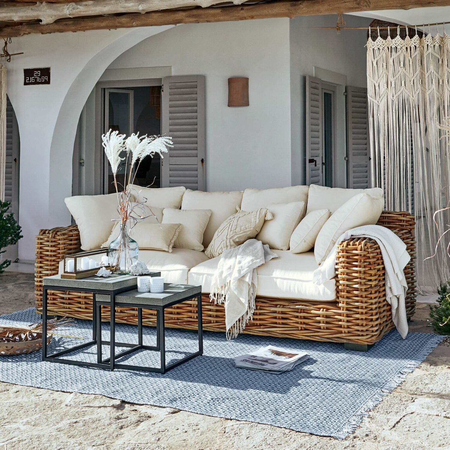 Sofa Rimini Sofá de ratán con cojines crema y manta sobre alfombra azul en terraza soleada