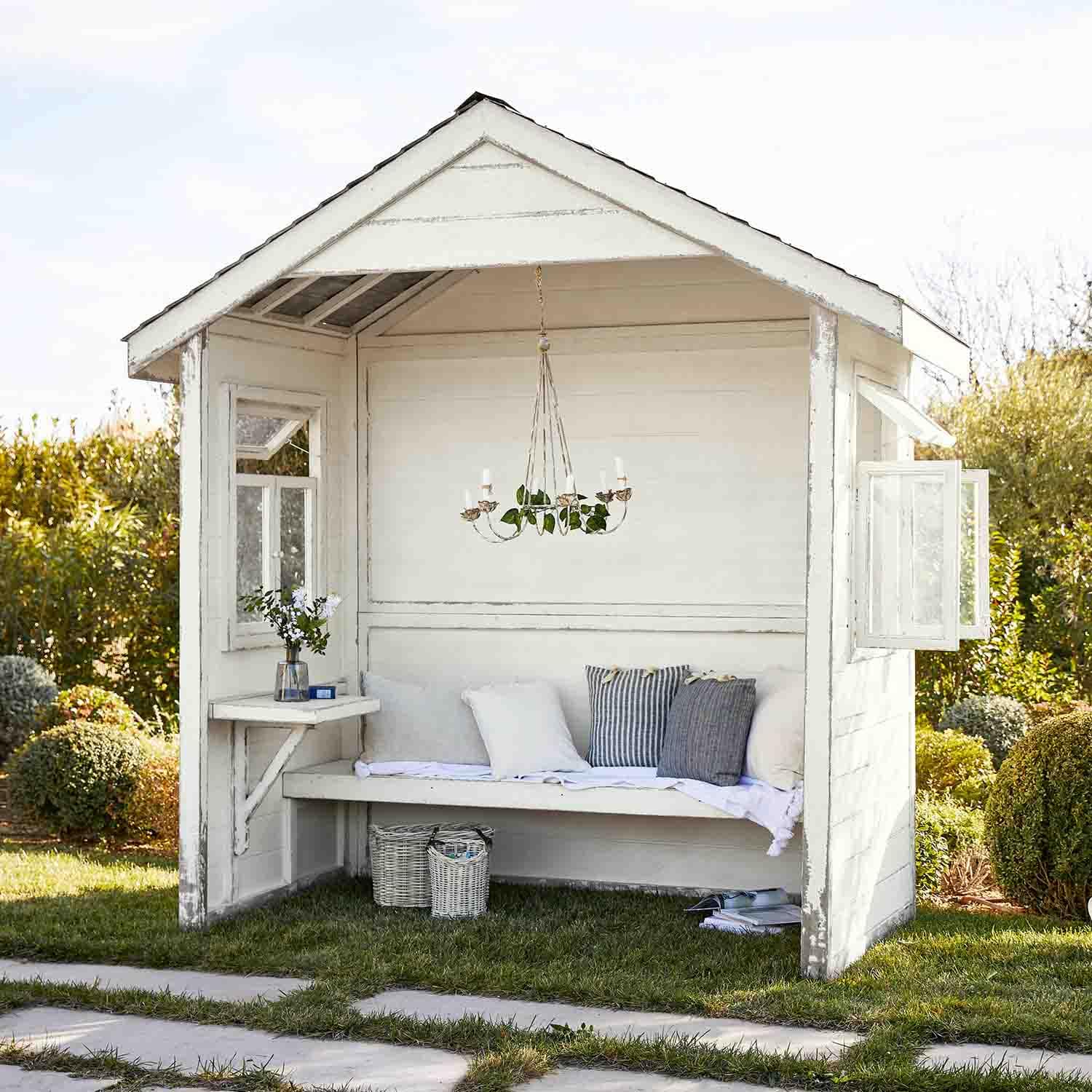 Pérgola de madera blanca con banco, cojines y candelabro colgante, espacio de relax en jardín