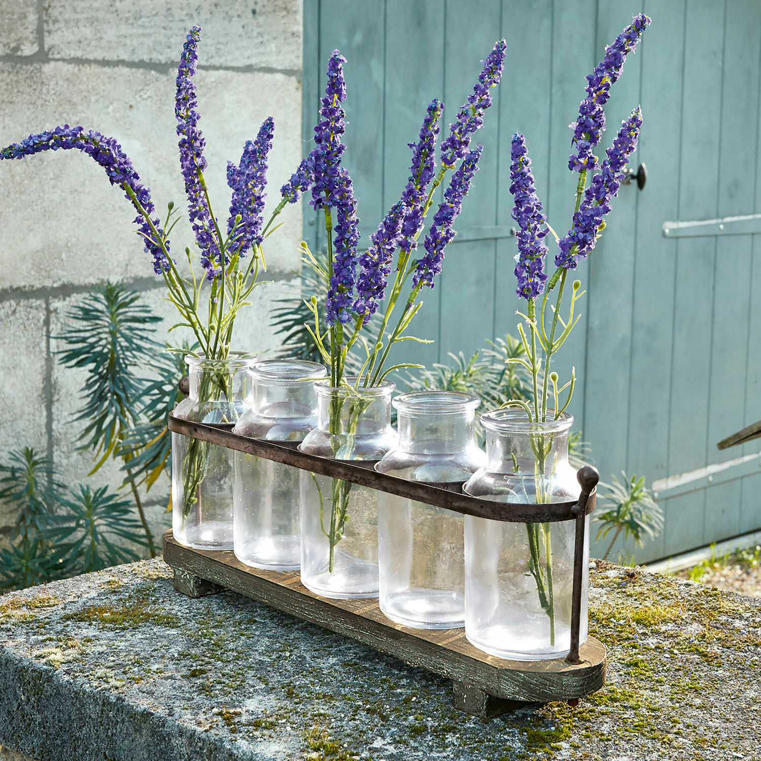 Set de 5 jarrones Ranjo Conjunto de cinco jarrones de vidrio transparente con soporte metálico sobre base de madera con flores moradas