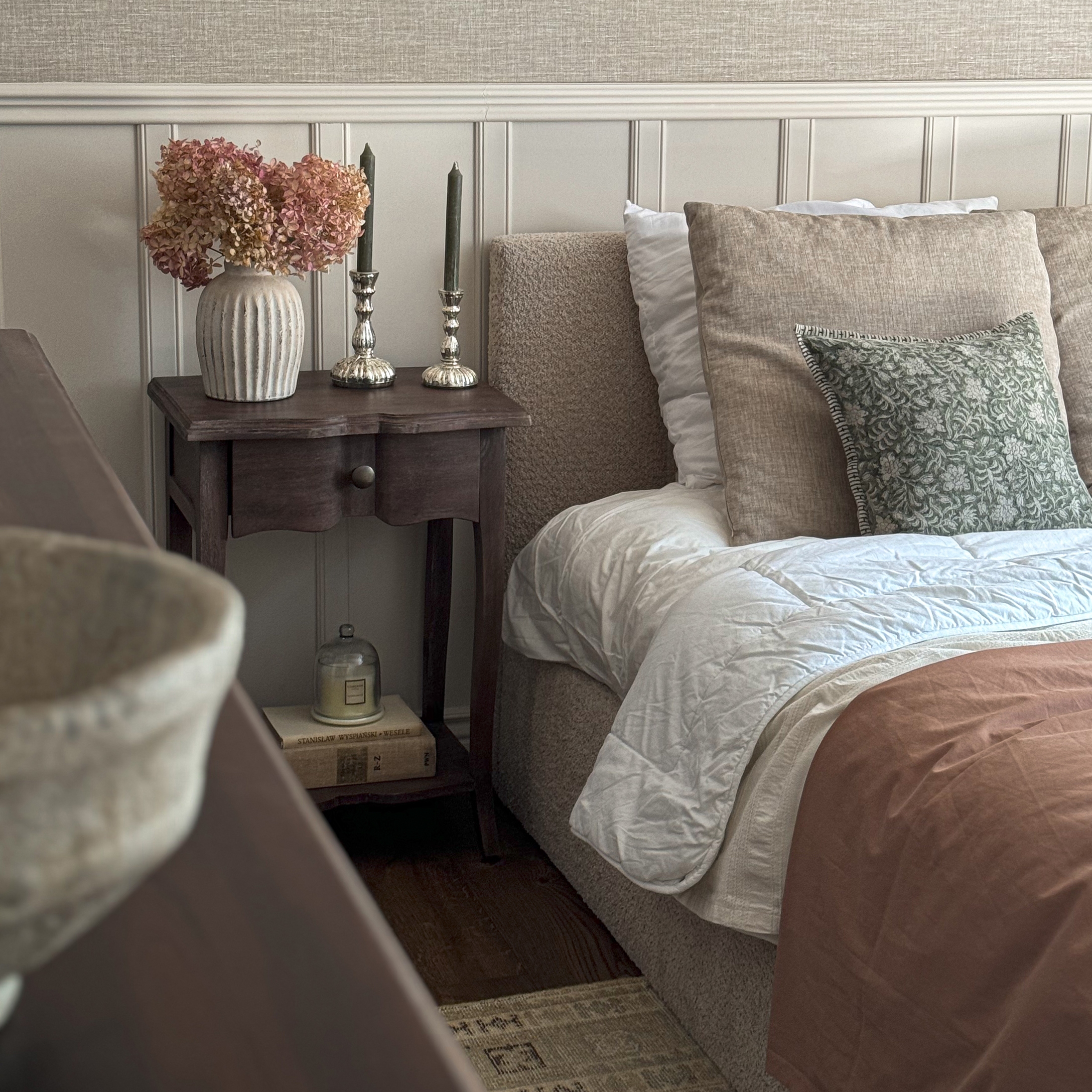 Dormitorio acogedor con ropa de cama beige, mesita de noche de madera y jarrón decorativo con flores secas