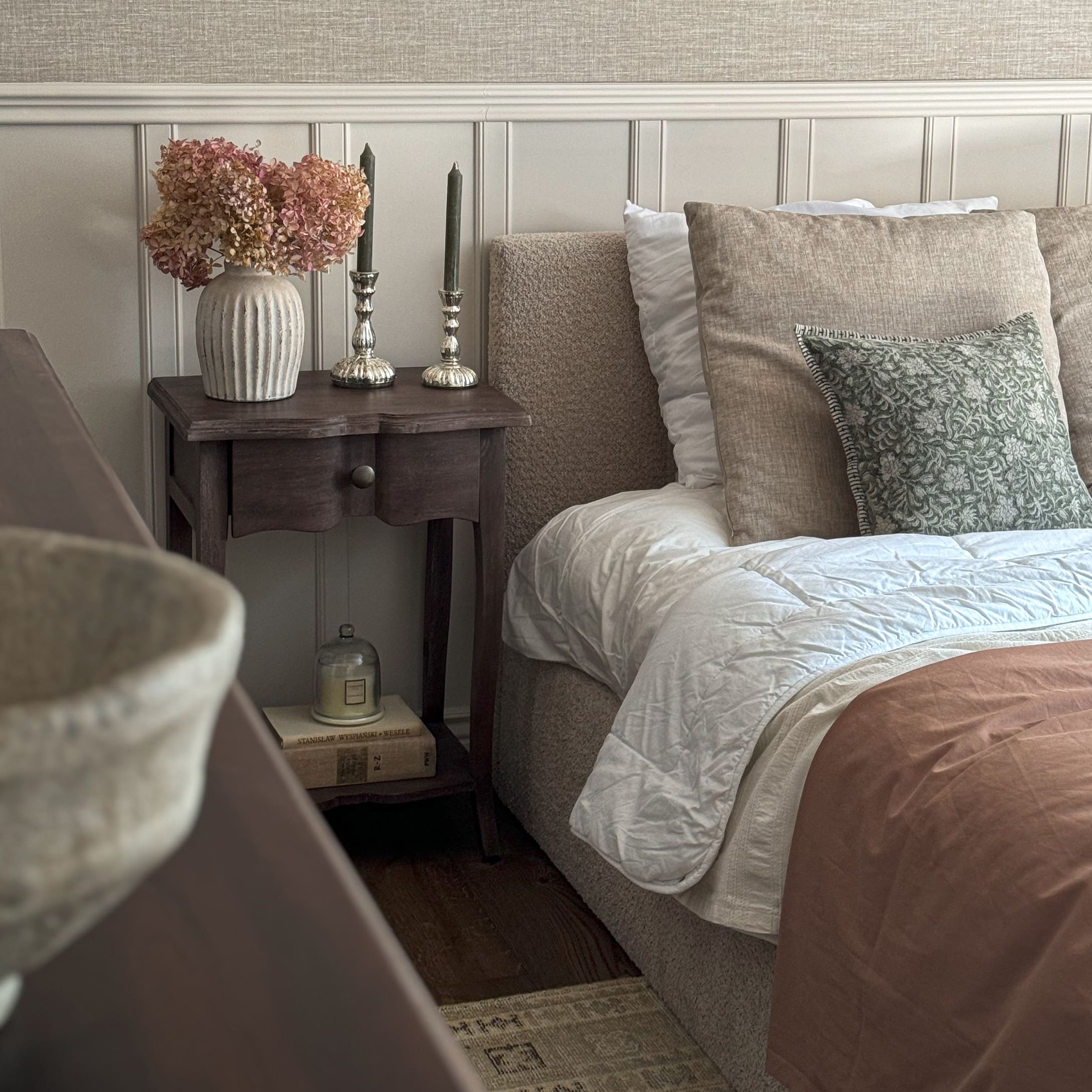 Dormitorio acogedor con ropa de cama beige, mesita de noche de madera y jarrón decorativo con flores secas