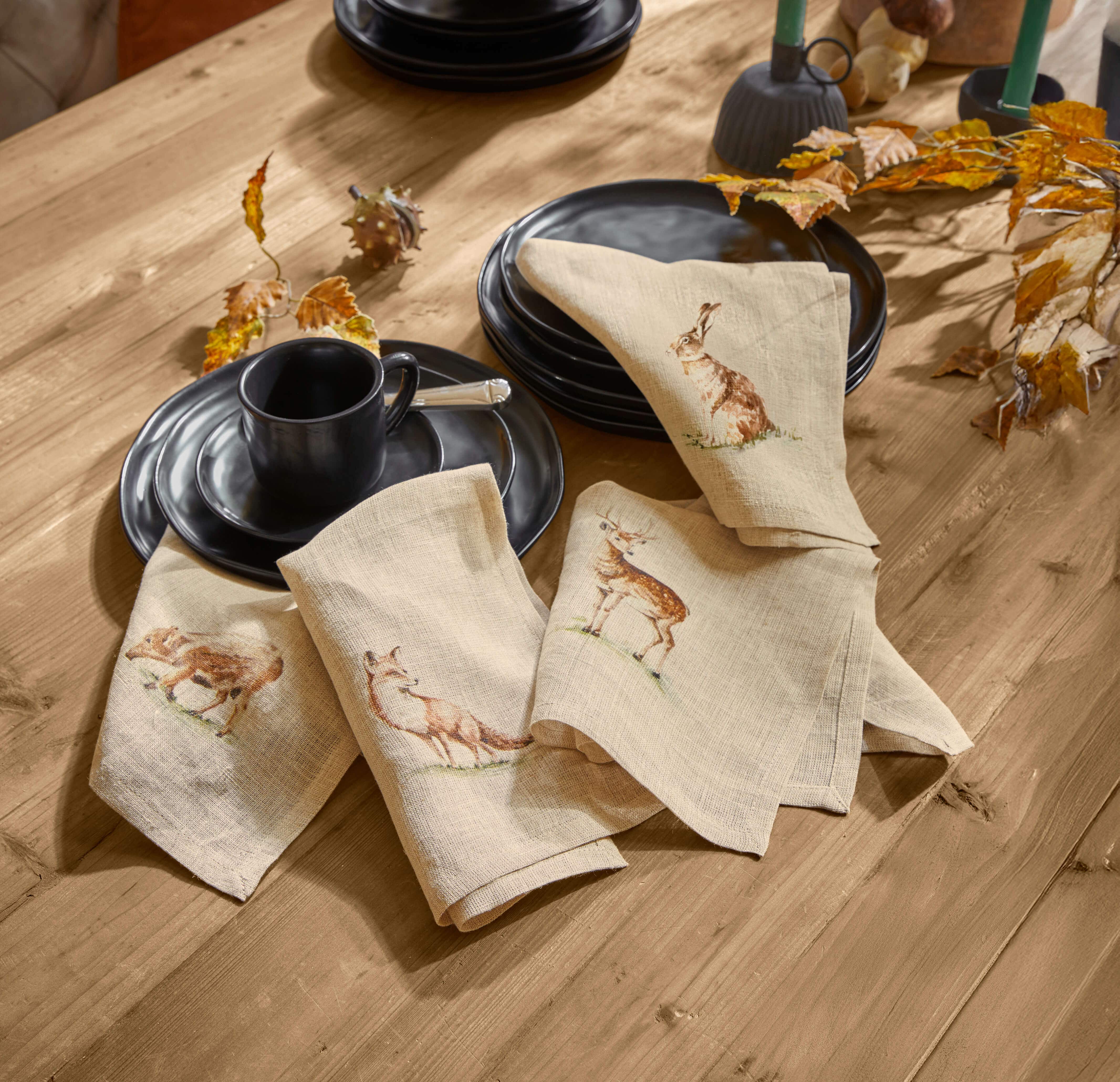 Servilletas de lino beige con estampados de animales del bosque sobre mesa de madera con vajilla negra