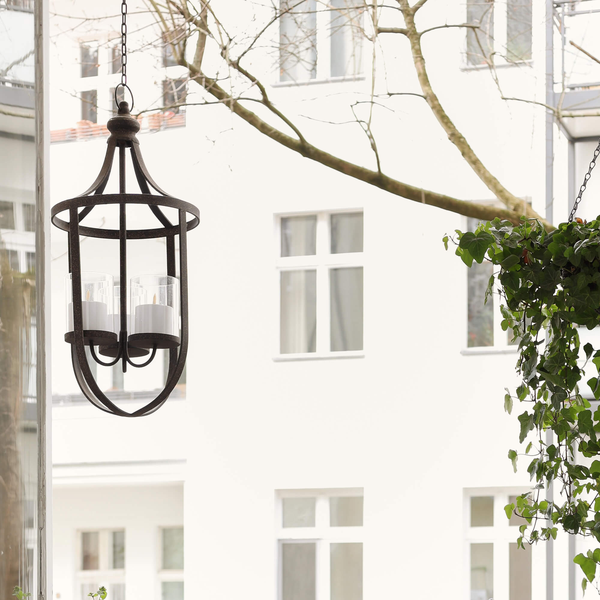 Farol colgante de metal con tres velas blancas frente a ventanas y plantas verdes en balcón