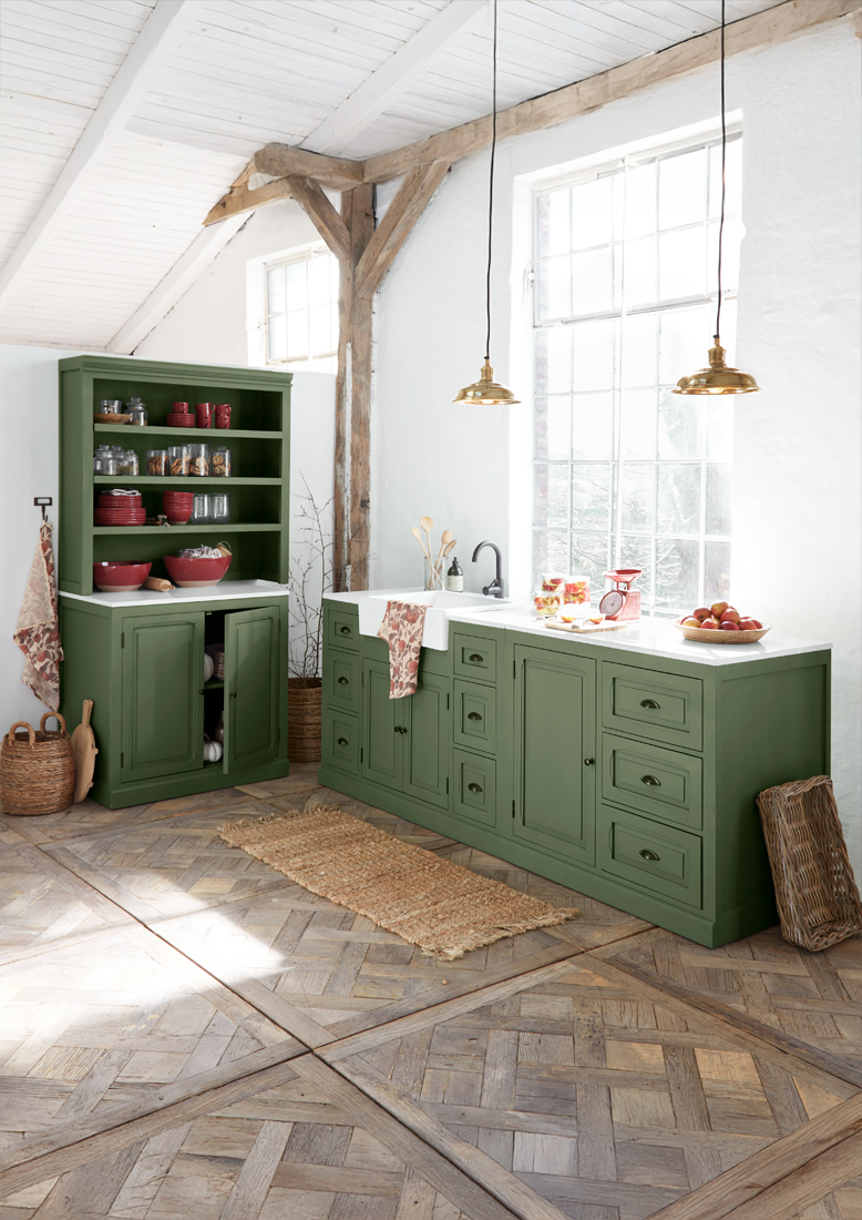 Cocina de madera verde con encimera blanca y lámparas de latón en interior rústico y luminoso