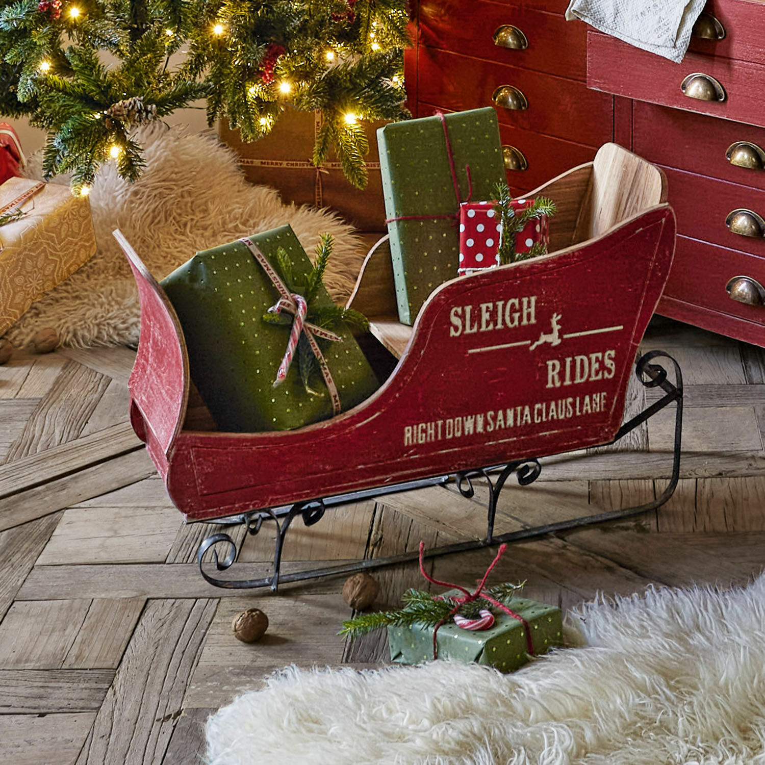 Trineo decorativo Maélys Trineo de madera rojo con patines metálicos y regalos verdes sobre suelo de madera junto a árbol de Navidad