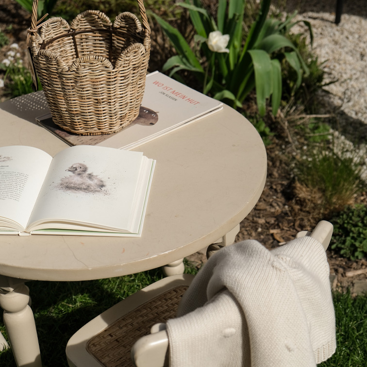 Mesa redonda beige de jardín con cesta de mimbre y libros abiertos sobre césped al sol