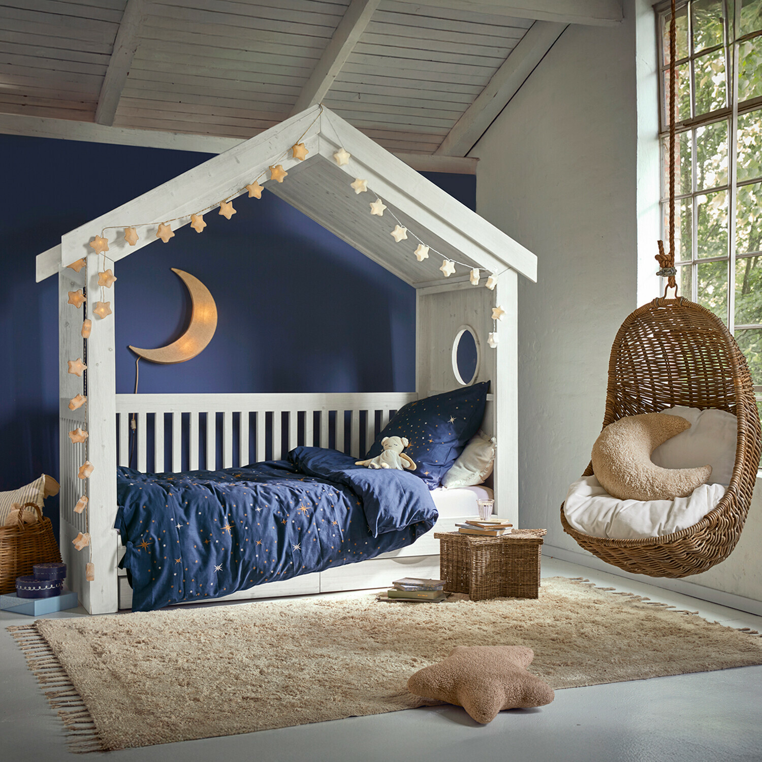 Alfombra Avenagh Cama de madera blanca con forma de casita, ropa de cama azul con estrellas y sillón colgante de mimbre en habitación infantil