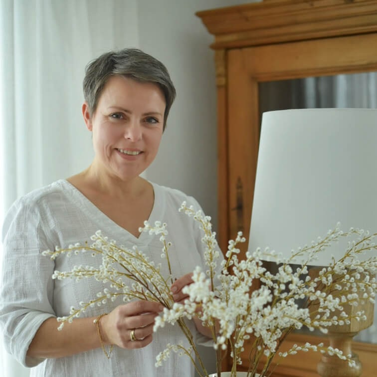 Mujer con blusa blanca sosteniendo ramas decorativas con flores blancas en interior luminoso