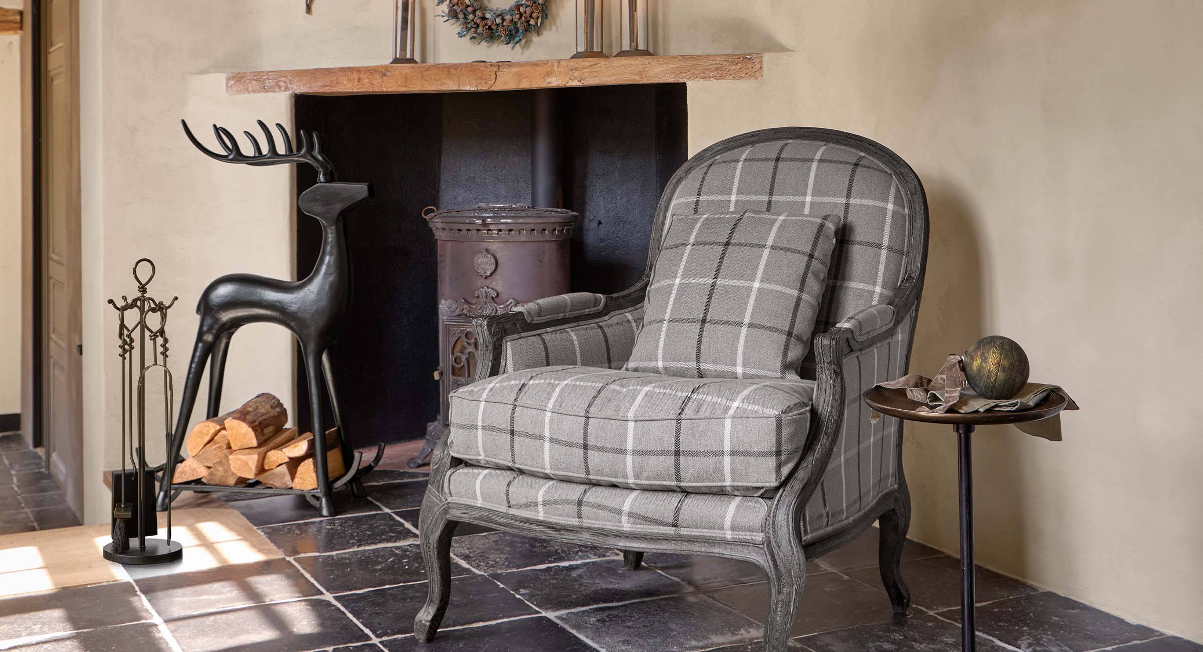 Sillón gris a cuadros con marco de madera junto a chimenea con soporte metálico para leña y herramientas