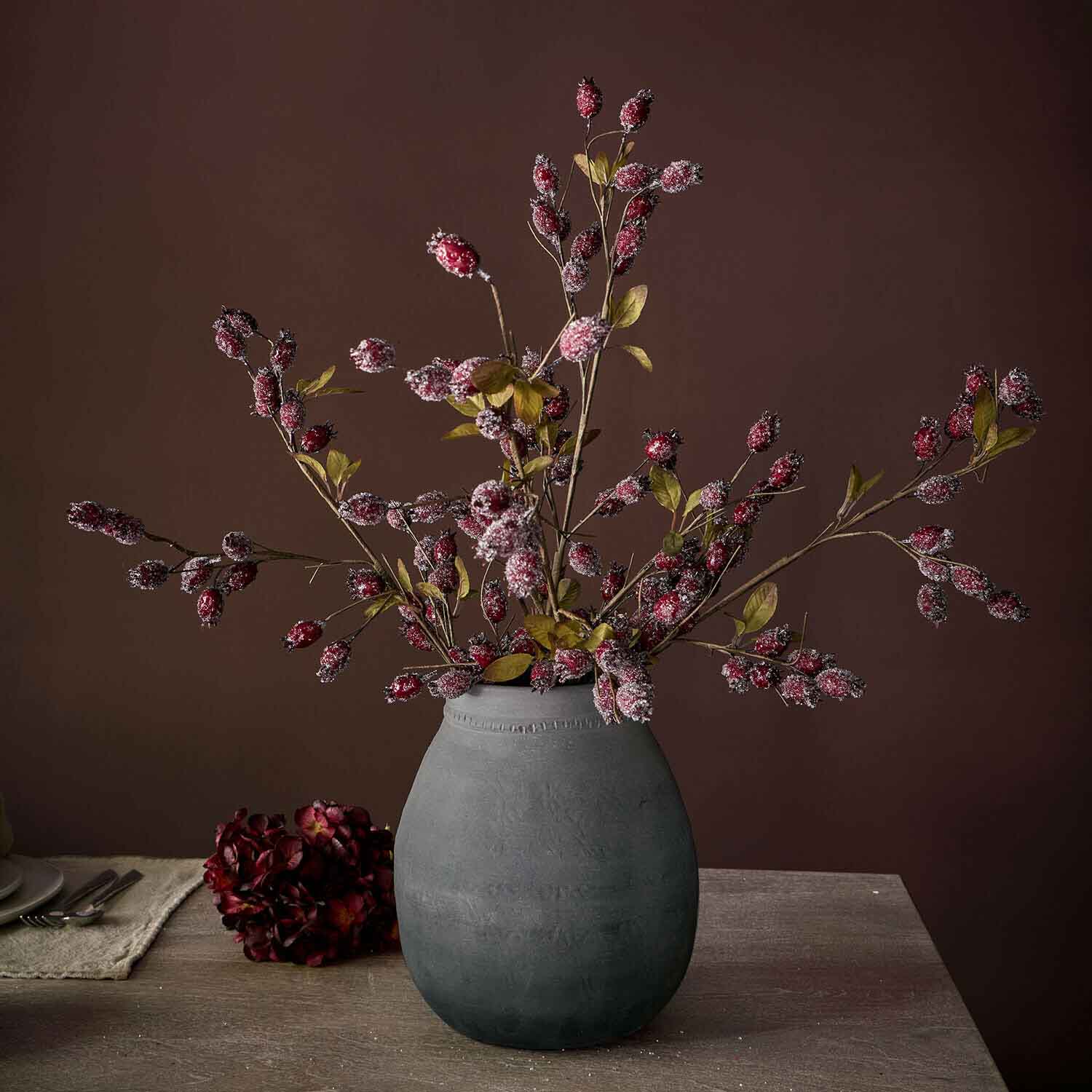 Jarrón de cerámica gris con ramas decorativas con frutos rojos y hojas verdes sobre mesa de madera