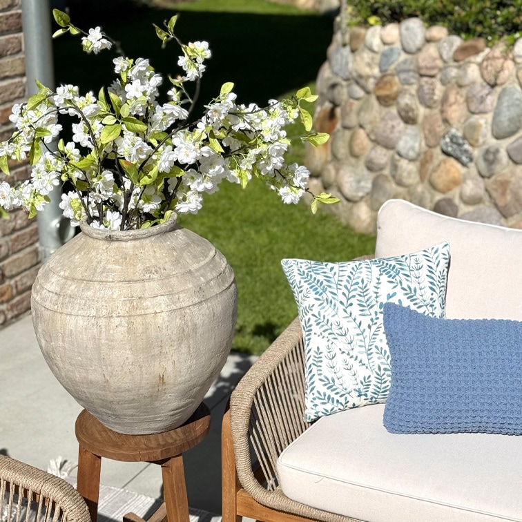 Gran jarrón de cerámica beige con flores blancas sobre soporte de madera junto a sofá con cojines en jardín