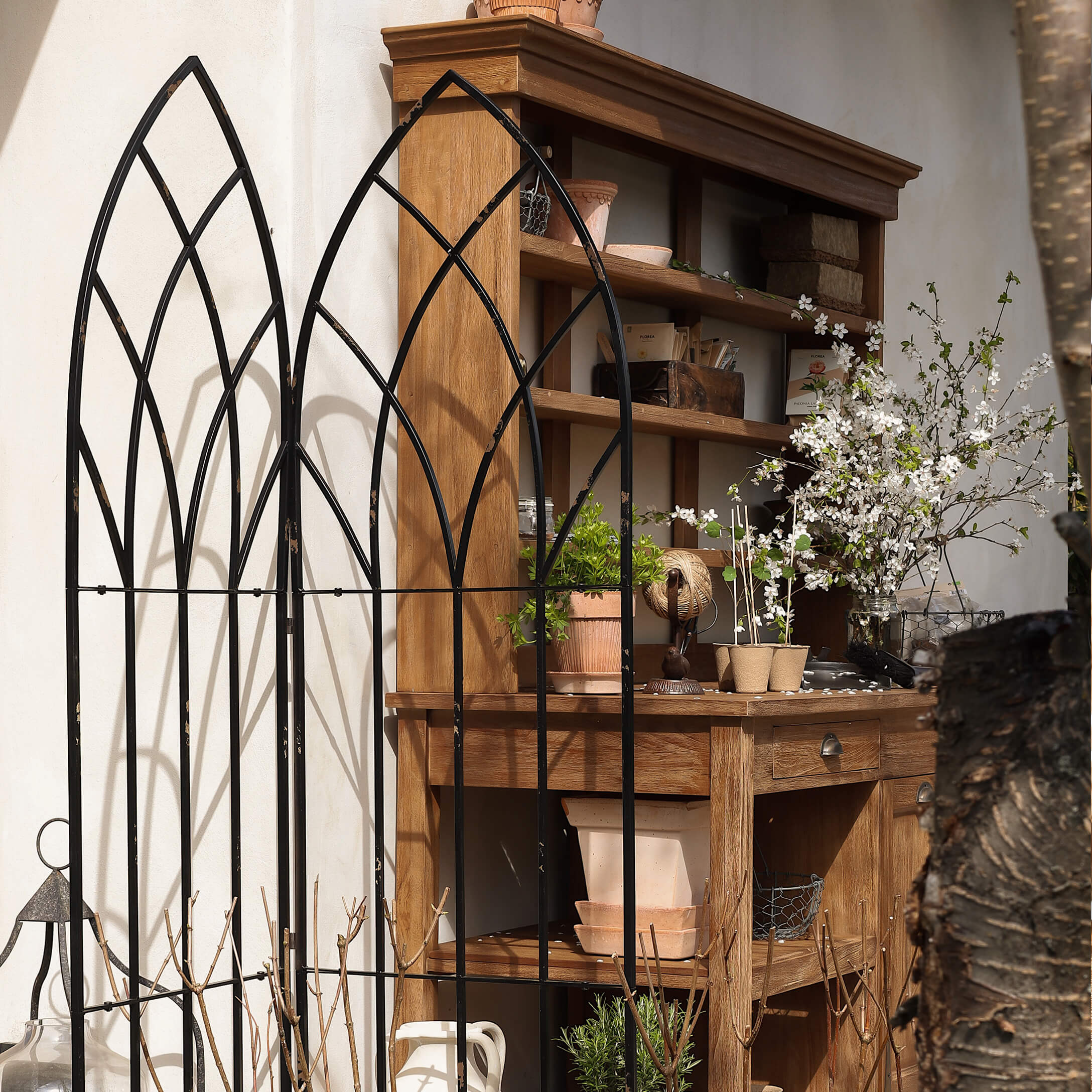 Reja de jardín de metal negro con diseño arqueado frente a estantería de madera con macetas y flores