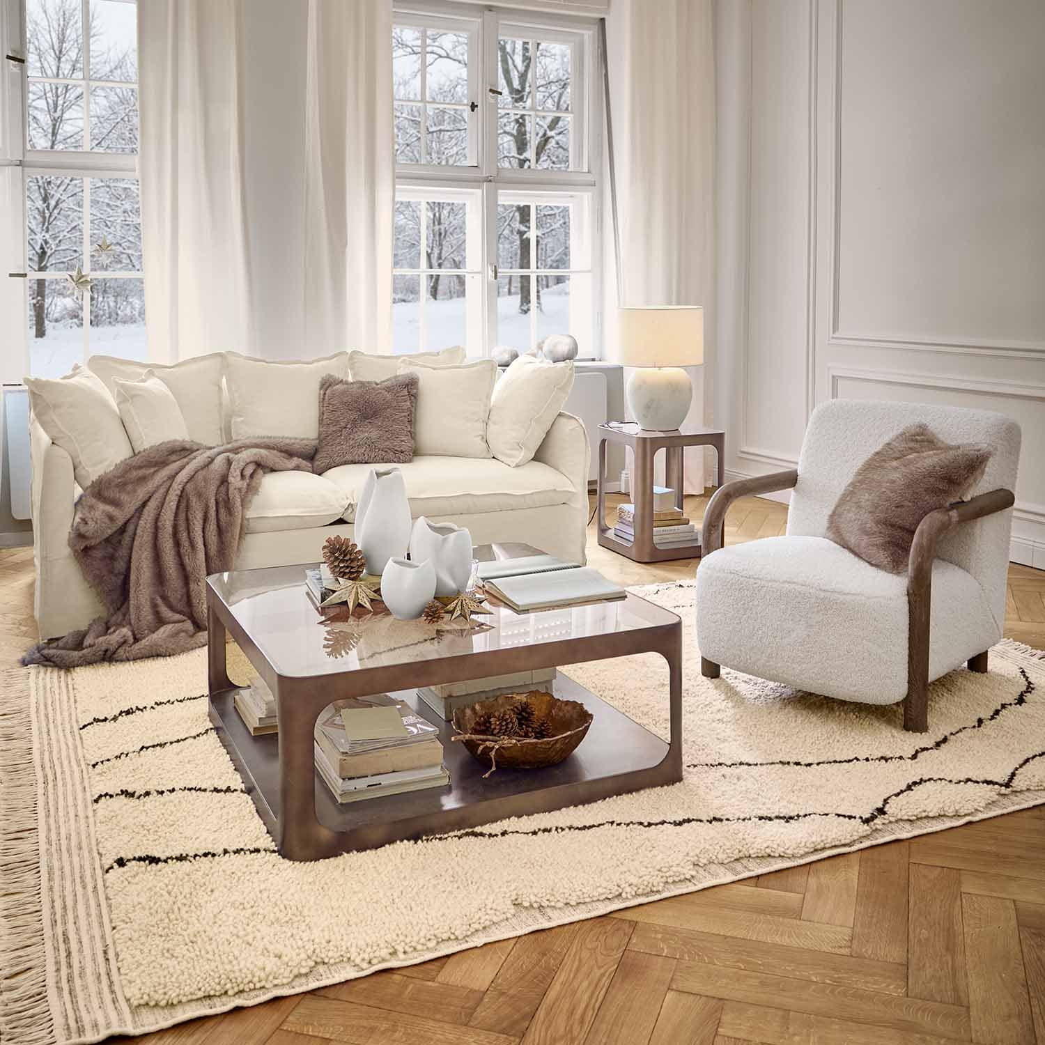 Sala acogedora con sofá blanco, sillón con reposabrazos de madera y alfombra suave en ambiente invernal