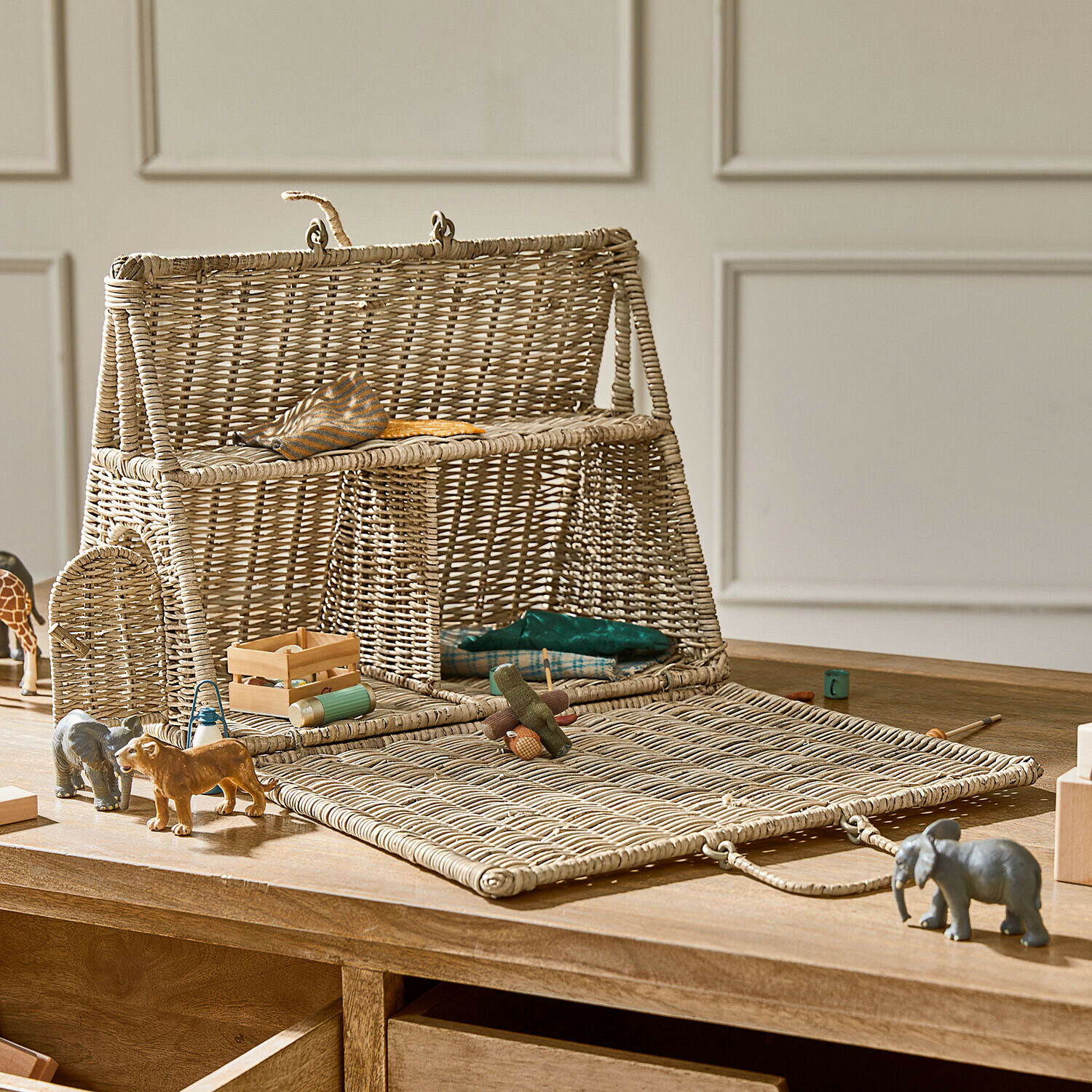 Casa de muñecas de mimbre natural con frente abatible y juguetes de madera sobre mesa