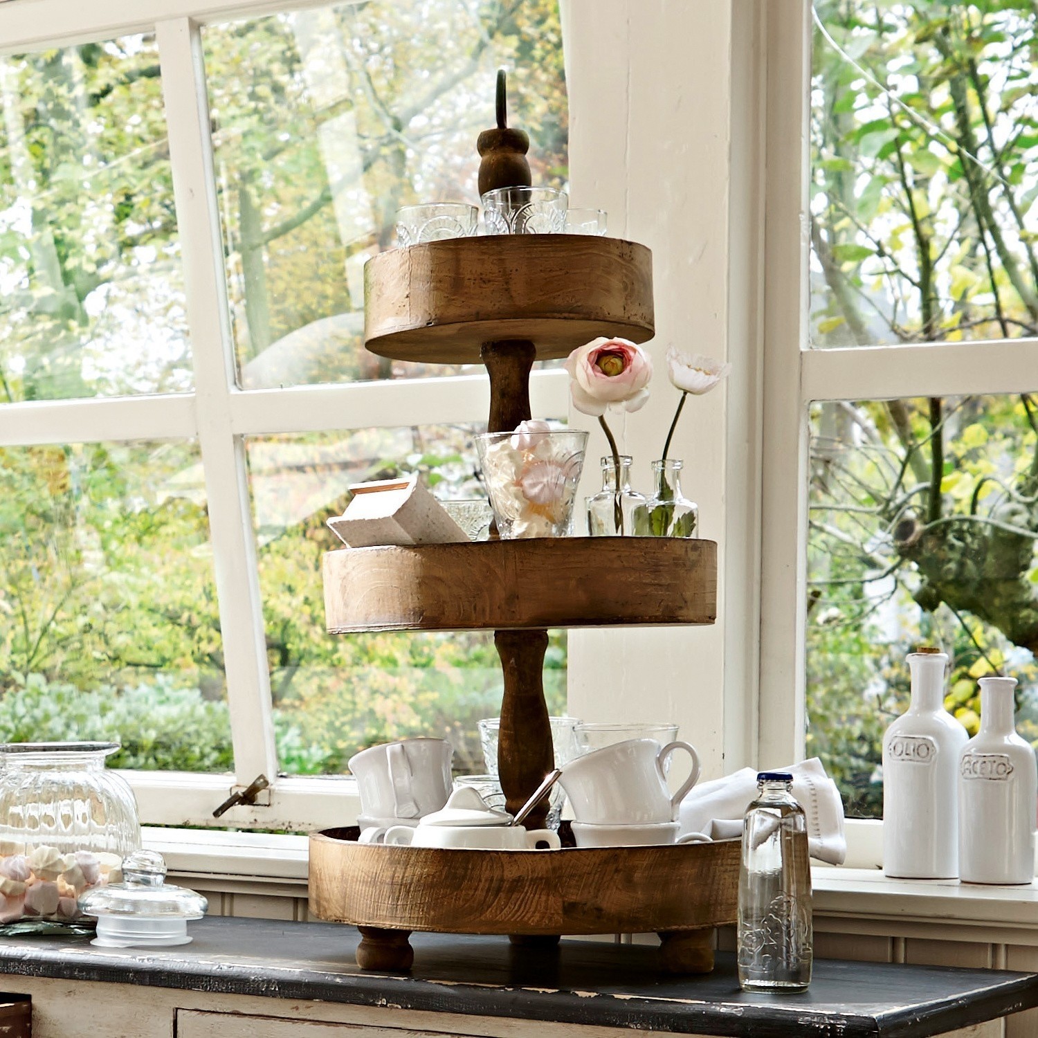 Estantería Beauvais Soporte de madera de tres niveles en color natural con tazas y decoraciones frente a ventana con vista a jardín