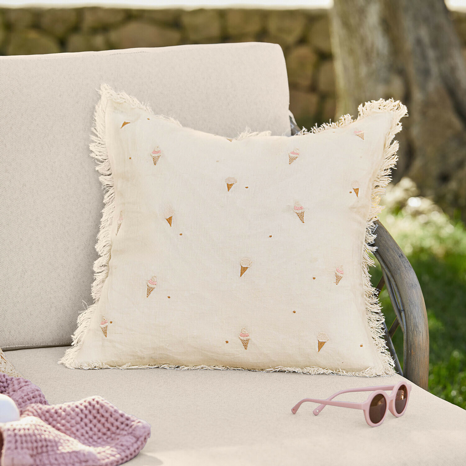 Cojín beige de lino con flecos y estampado de conos de helado sobre sofá claro en jardín