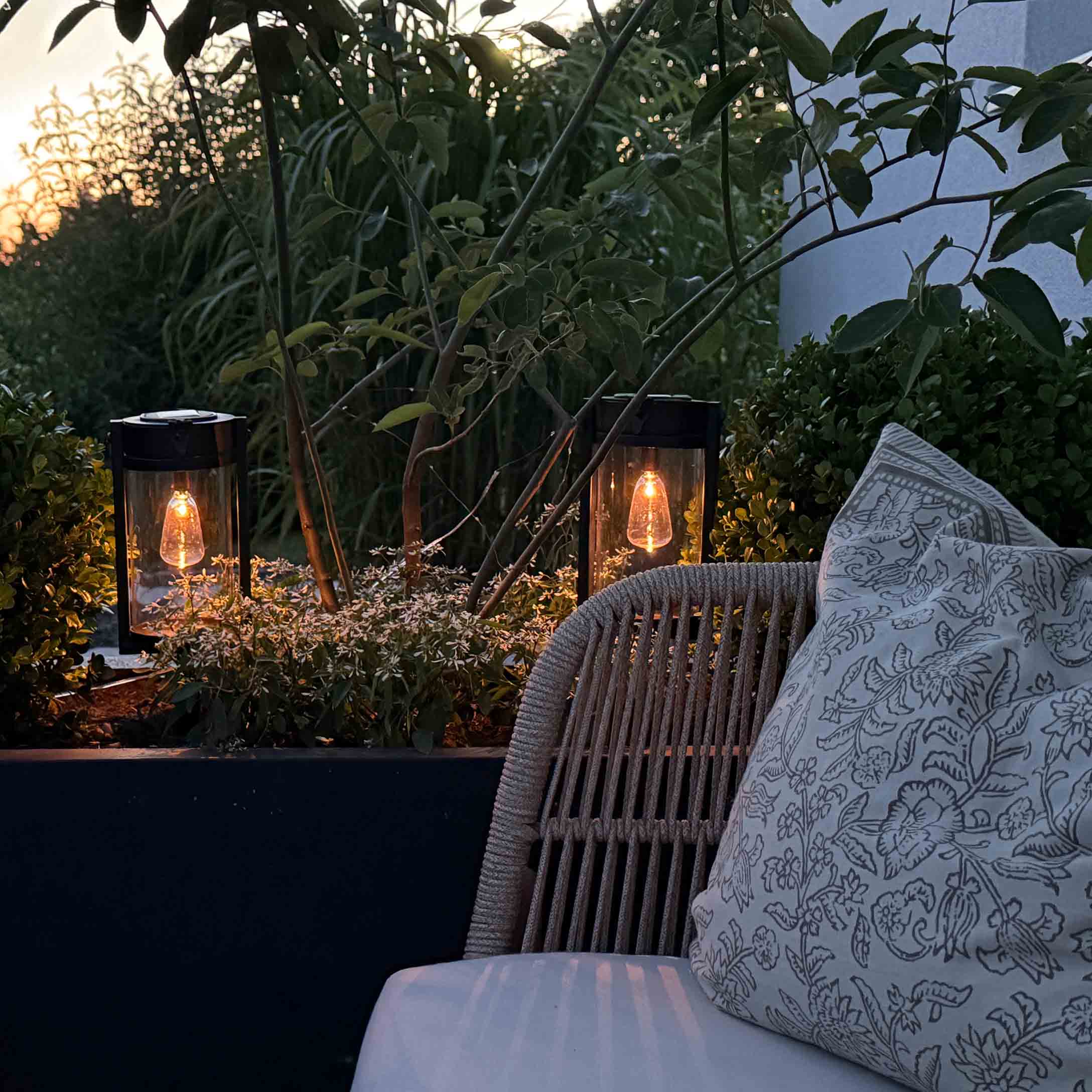 Faroles de jardín de metal negro con luz cálida entre plantas verdes en la terraza
