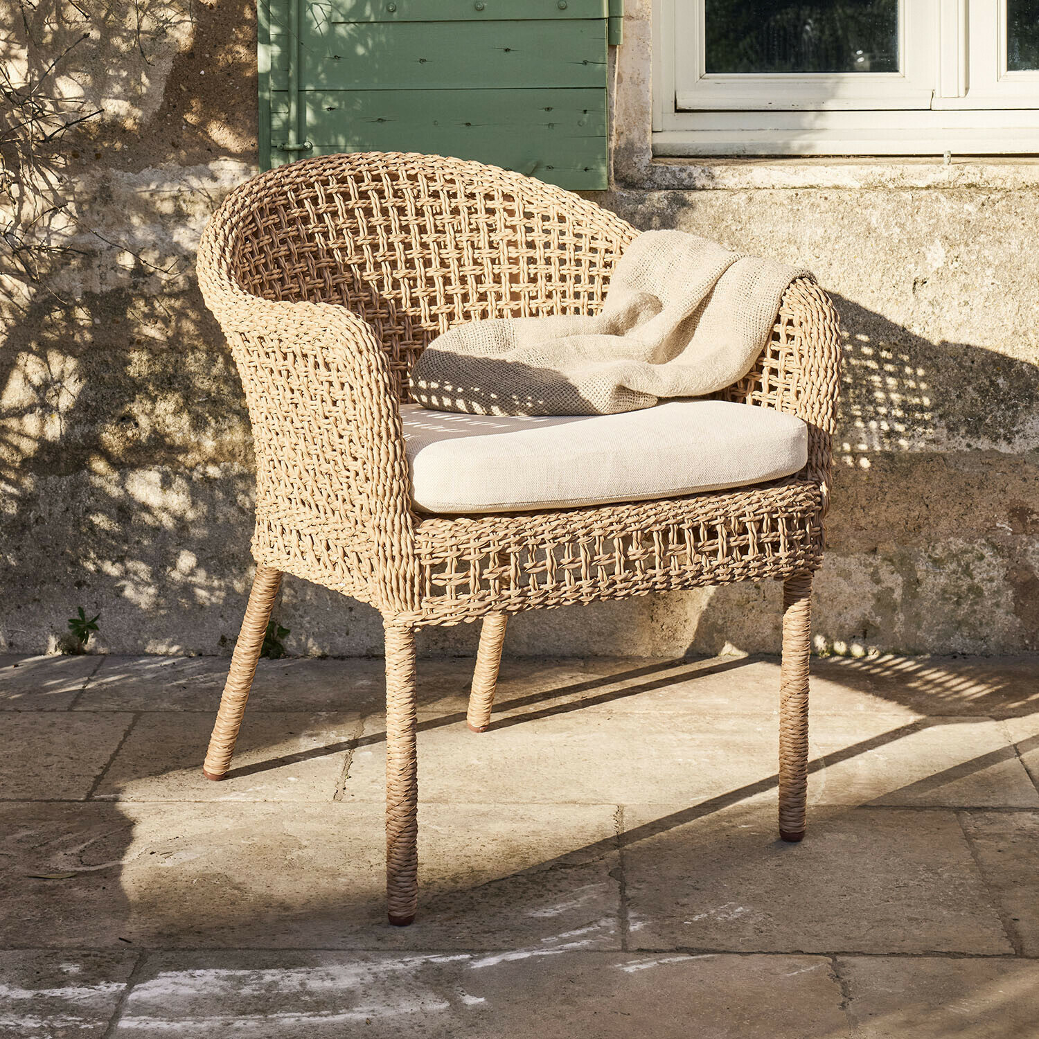 Silla Mossbury Sillón de ratán trenzado natural con cojín claro y manta beige en terraza de piedra