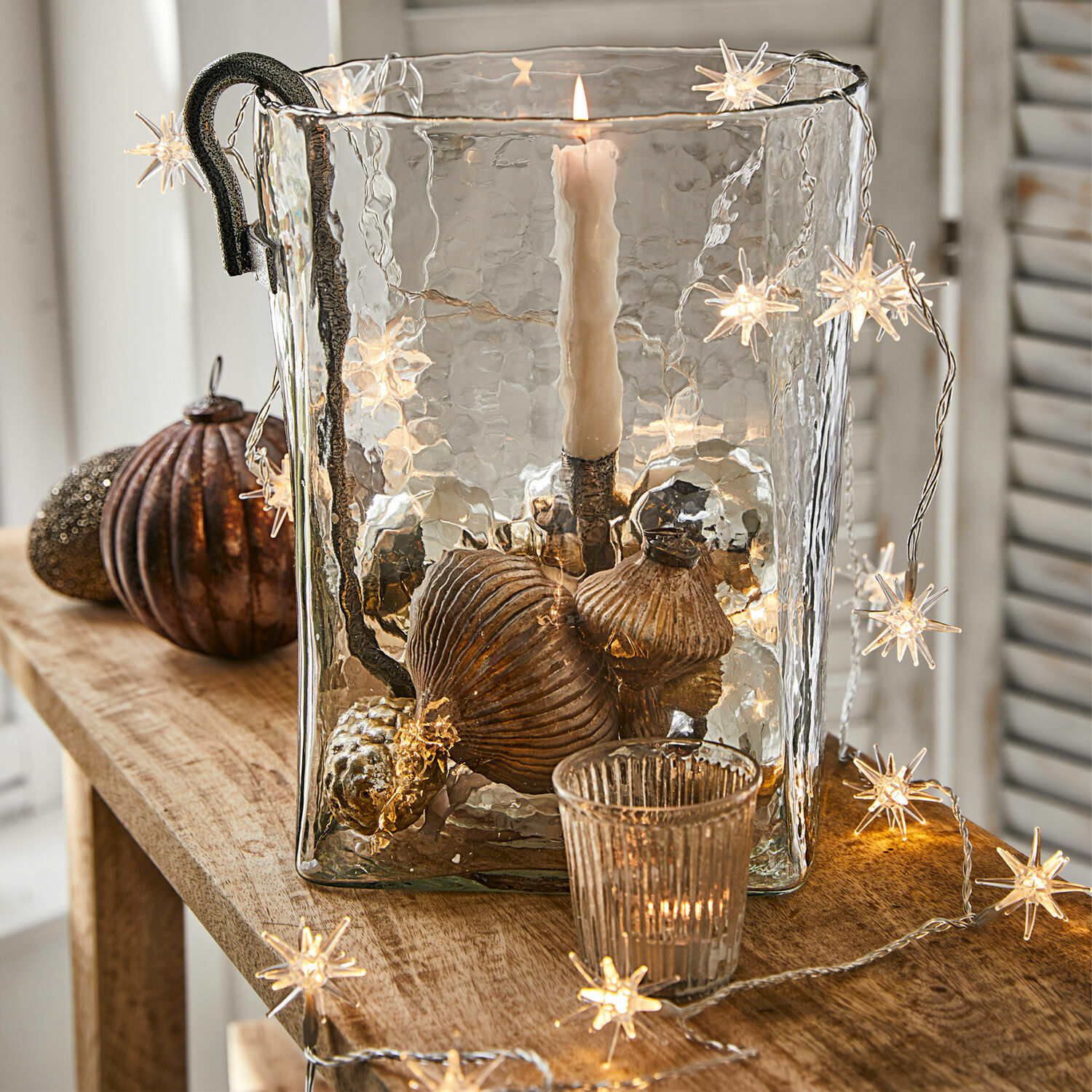 Luces de hadas Nelisse Portavelas de vidrio con asa metálica y decoraciones en cobre sobre mesa de madera con luces en forma de estrella