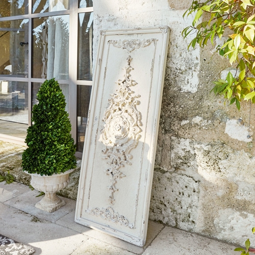Panel decorativo blanco de madera con relieve vintage apoyado en pared de piedra junto a planta verde