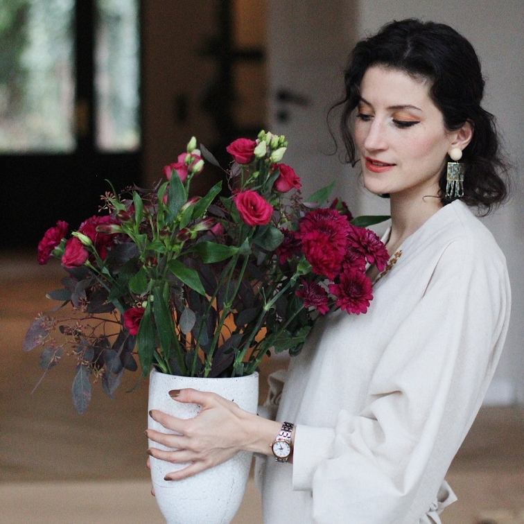 Mujer con blusa clara sosteniendo un jarrón de cerámica blanca con flores rojas en interior