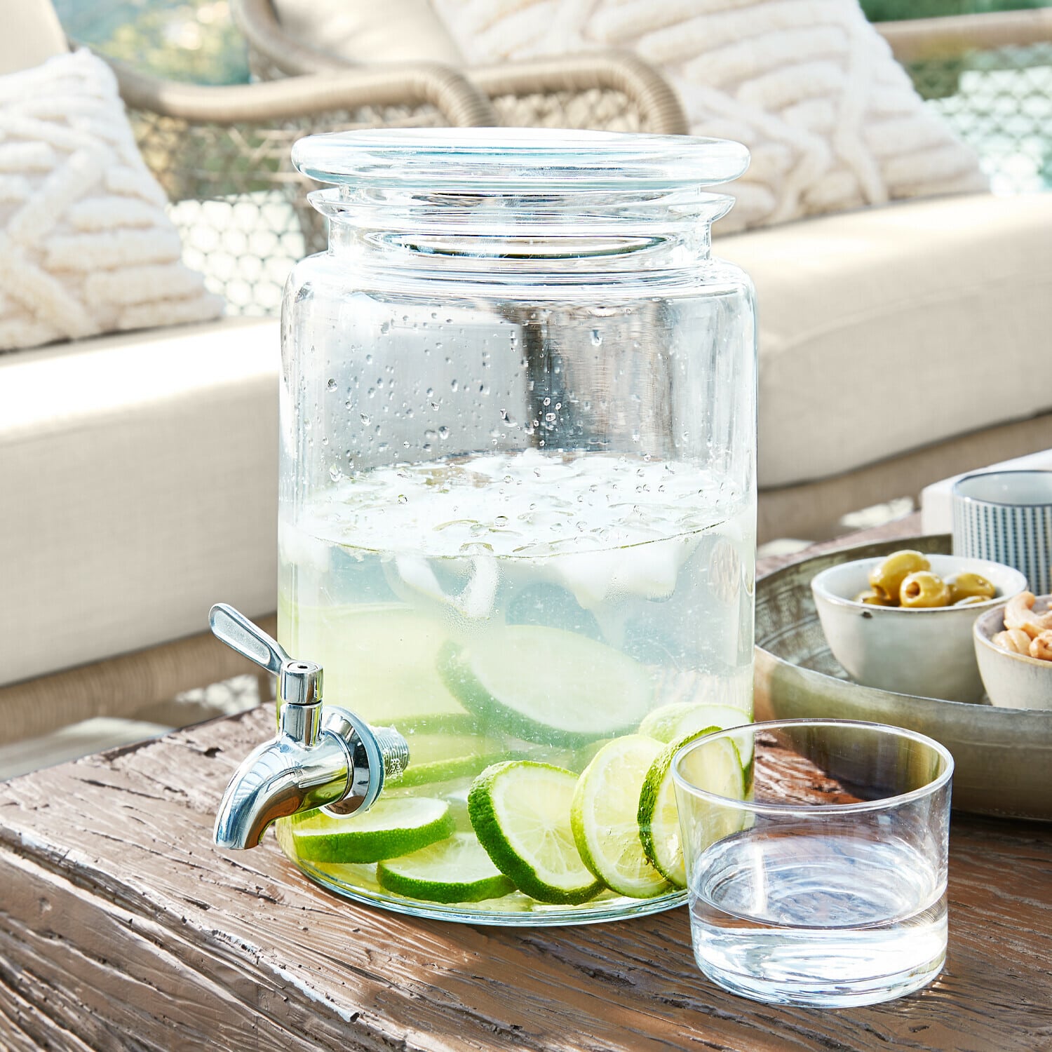 Dispensador de bebidas de vidrio con grifo, lleno de agua con rodajas de lima sobre mesa de madera