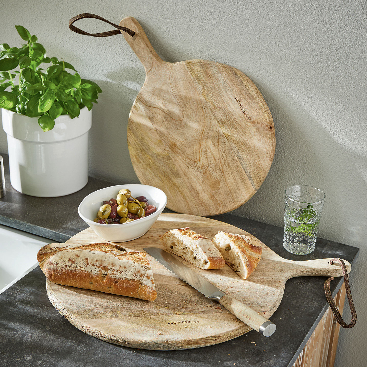 Juego de 2 tablas de cortar Nitra Tabla de cortar redonda de madera natural con mango, cuchillo de pan, pan cortado y aceitunas en encimera