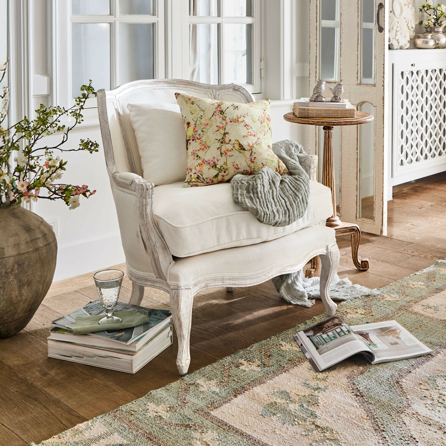 Sillón Kingswinford Sillón de madera blanca con tapicería clara, cojín floral y manta gris en salón acogedor